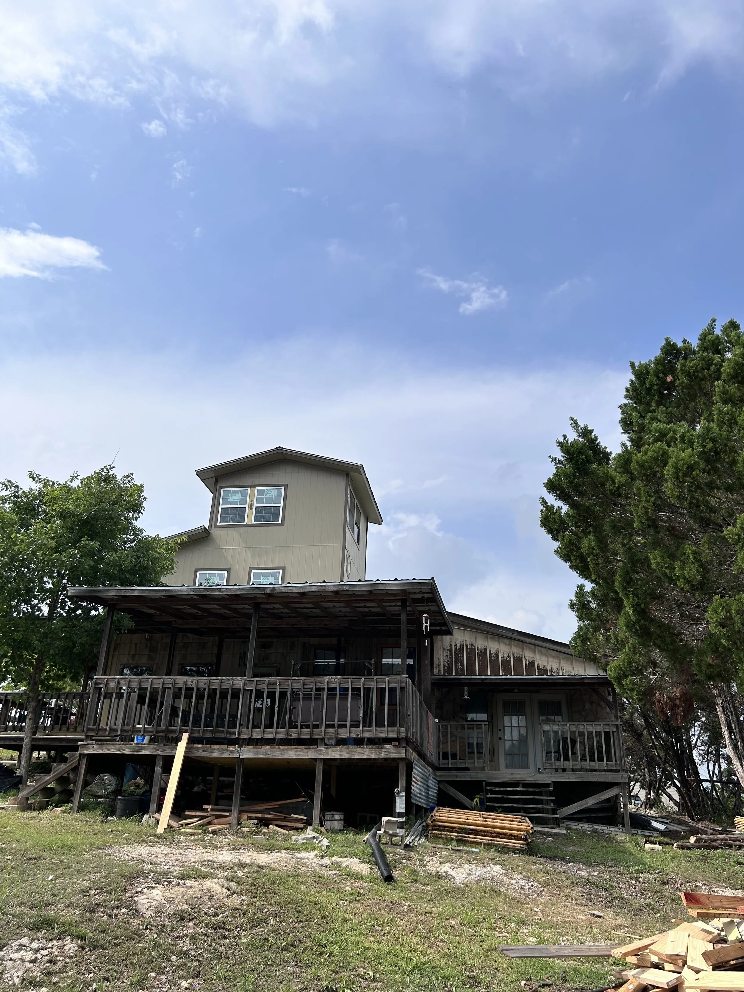A house with a raised wooden deck, stairs, and a smaller extension on top, surrounded by trees and a grassy yard with construction materials.