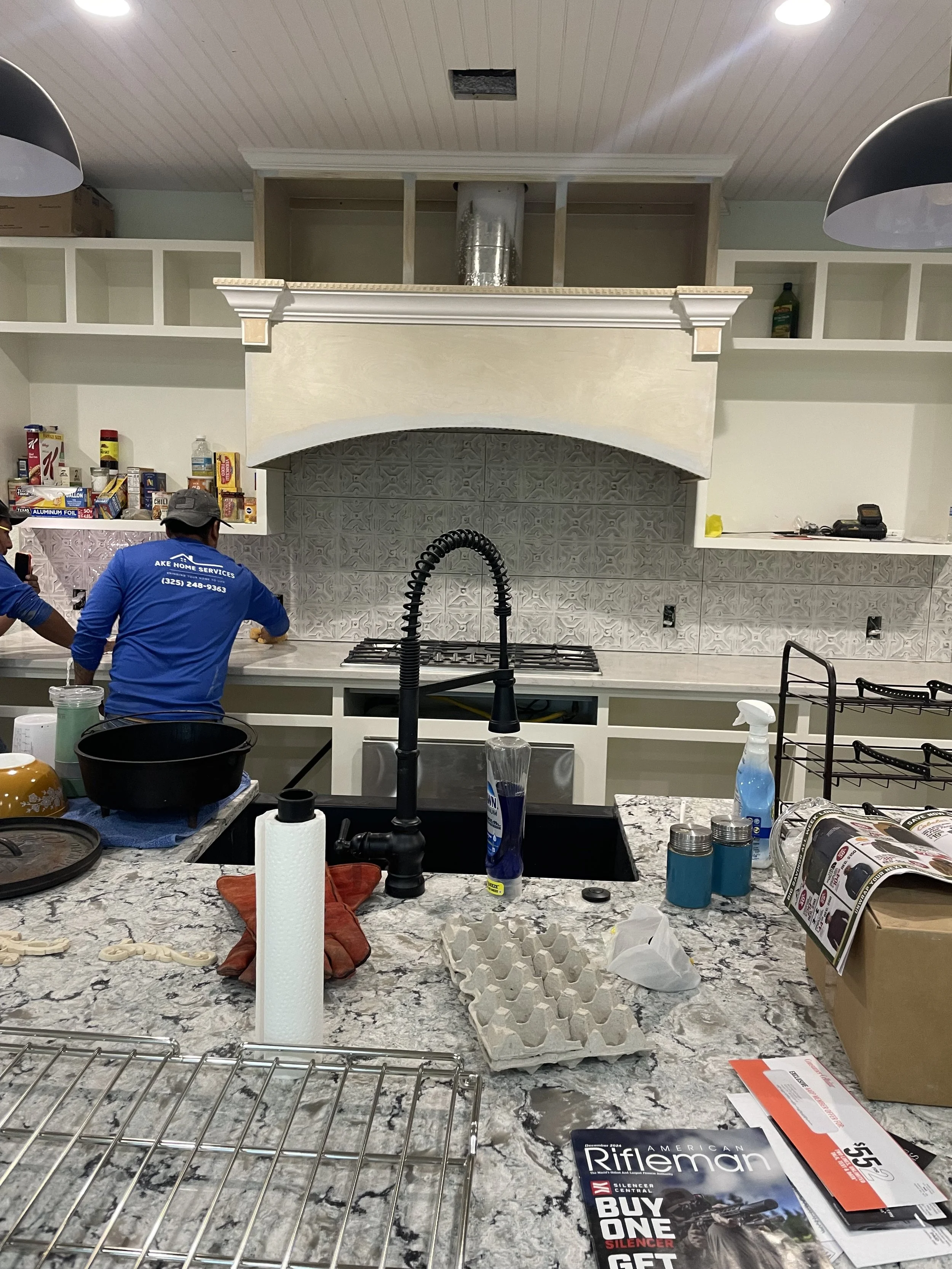 The image shows a cluttered kitchen island with various items including paper towels, a cardboard box, a magazine, small jars, a cleaning spray bottle, and egg cartons. In the background, two workers are installing or repairing cabinets above a stove