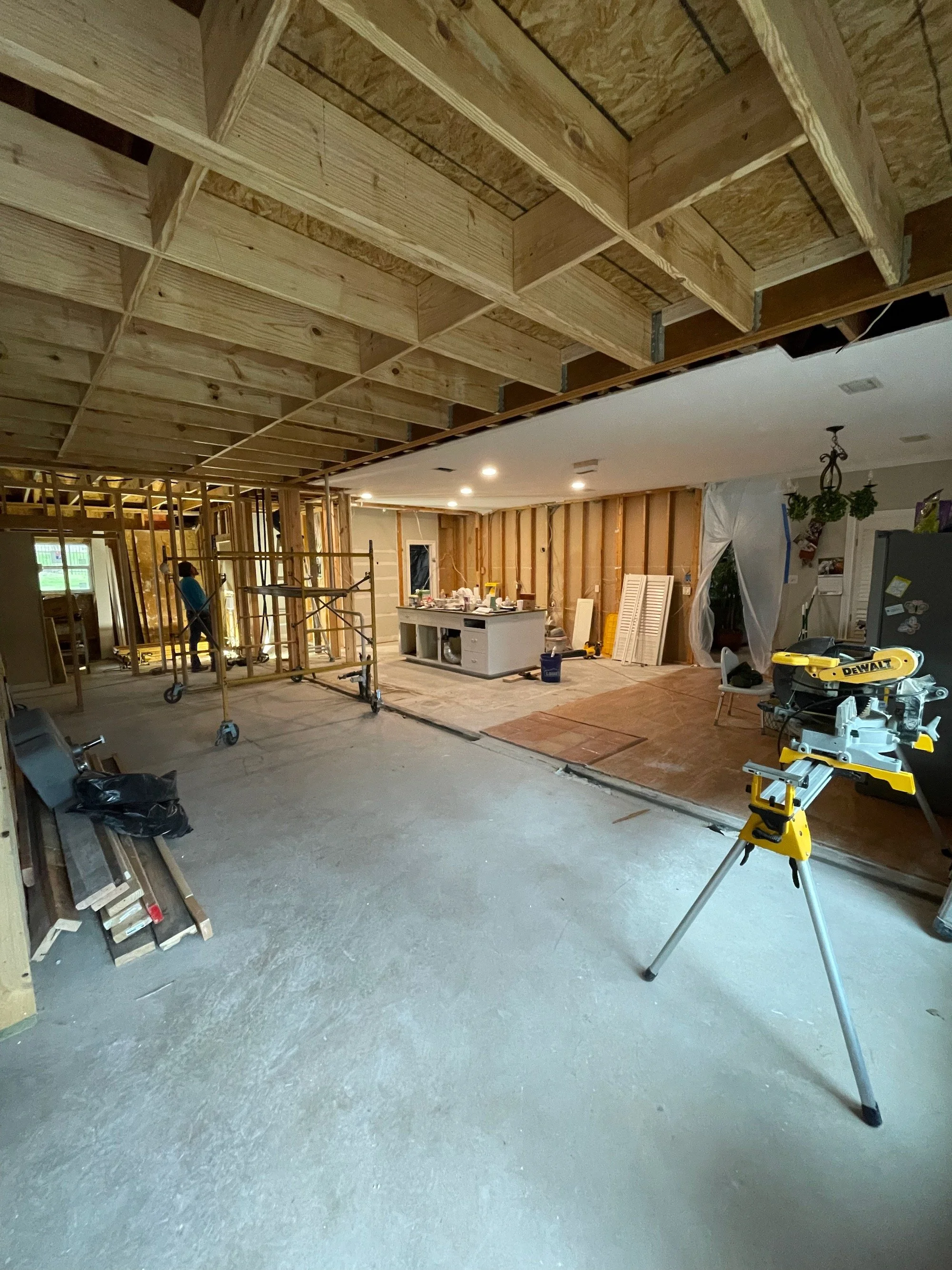 Home renovation site with exposed ceiling joists and wall framing, scaffolding, a kitchen island, construction tools, and a worker.