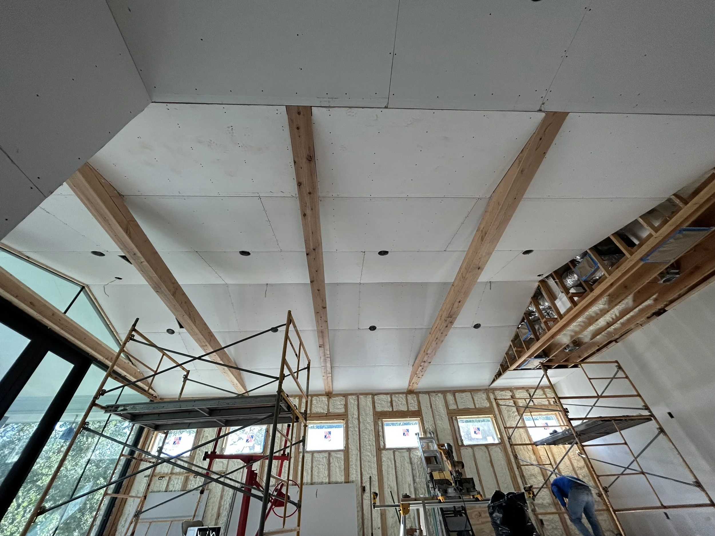 Construction workers installing a drywall ceiling with wooden beams, scaffolding, and tools inside a building under renovation.