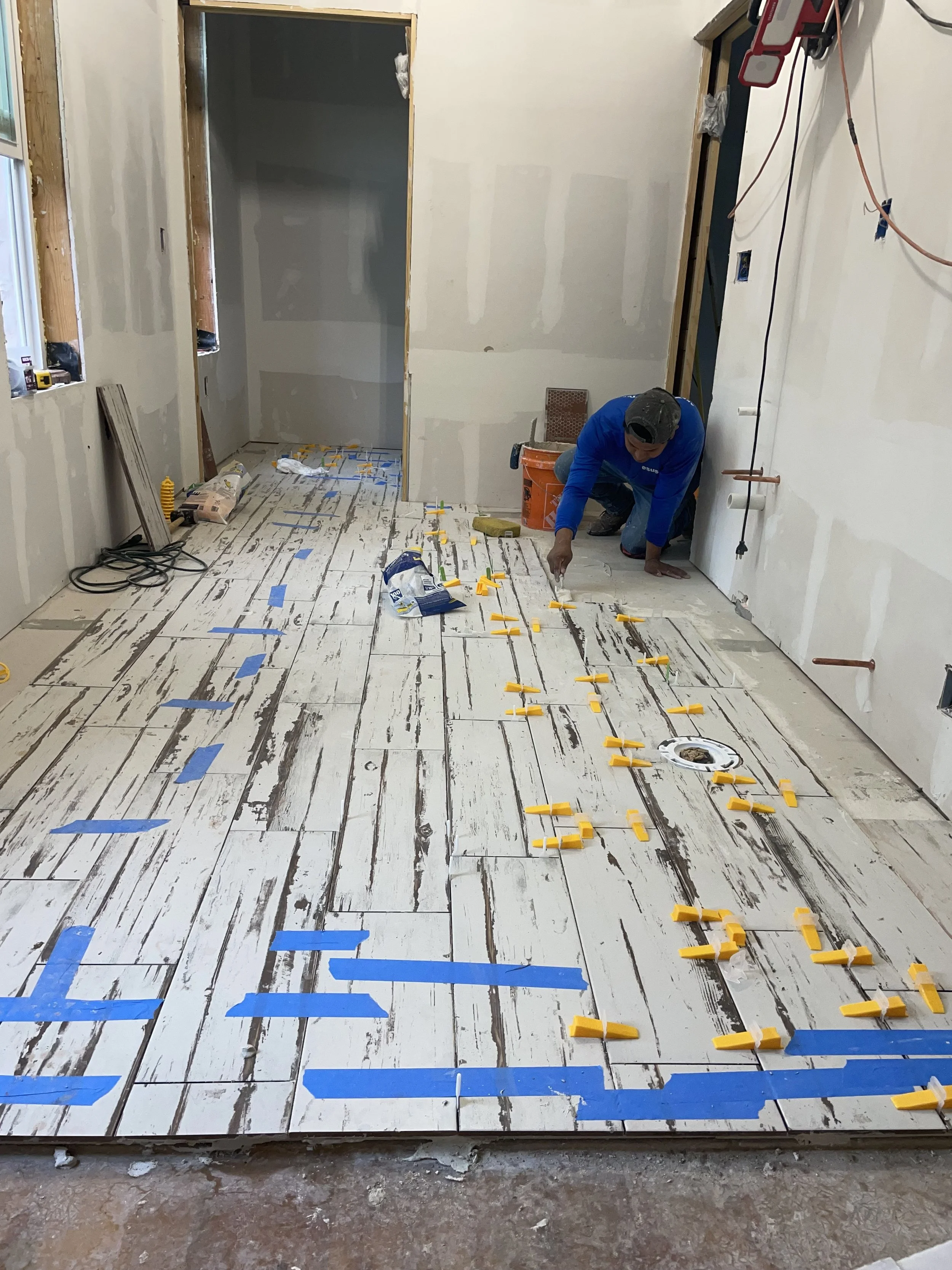 A construction worker installing hardwood flooring, using yellow spacers and blue painter's tape to align the planks. The room is under renovation with unfinished walls and exposed wiring.