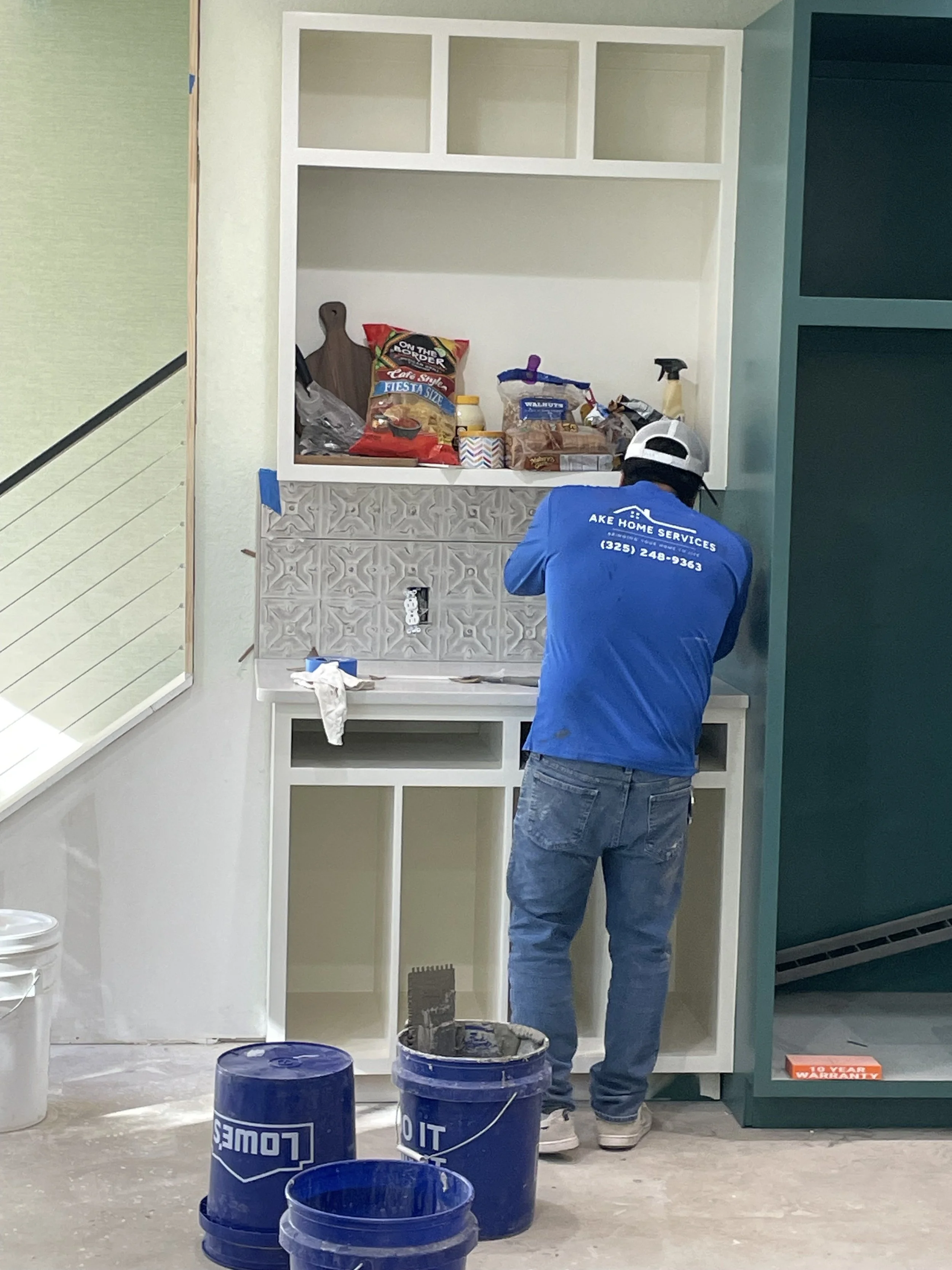 Worker installing a kitchen cabinet or countertop, surrounded by tools and materials, with blue buckets in the foreground.