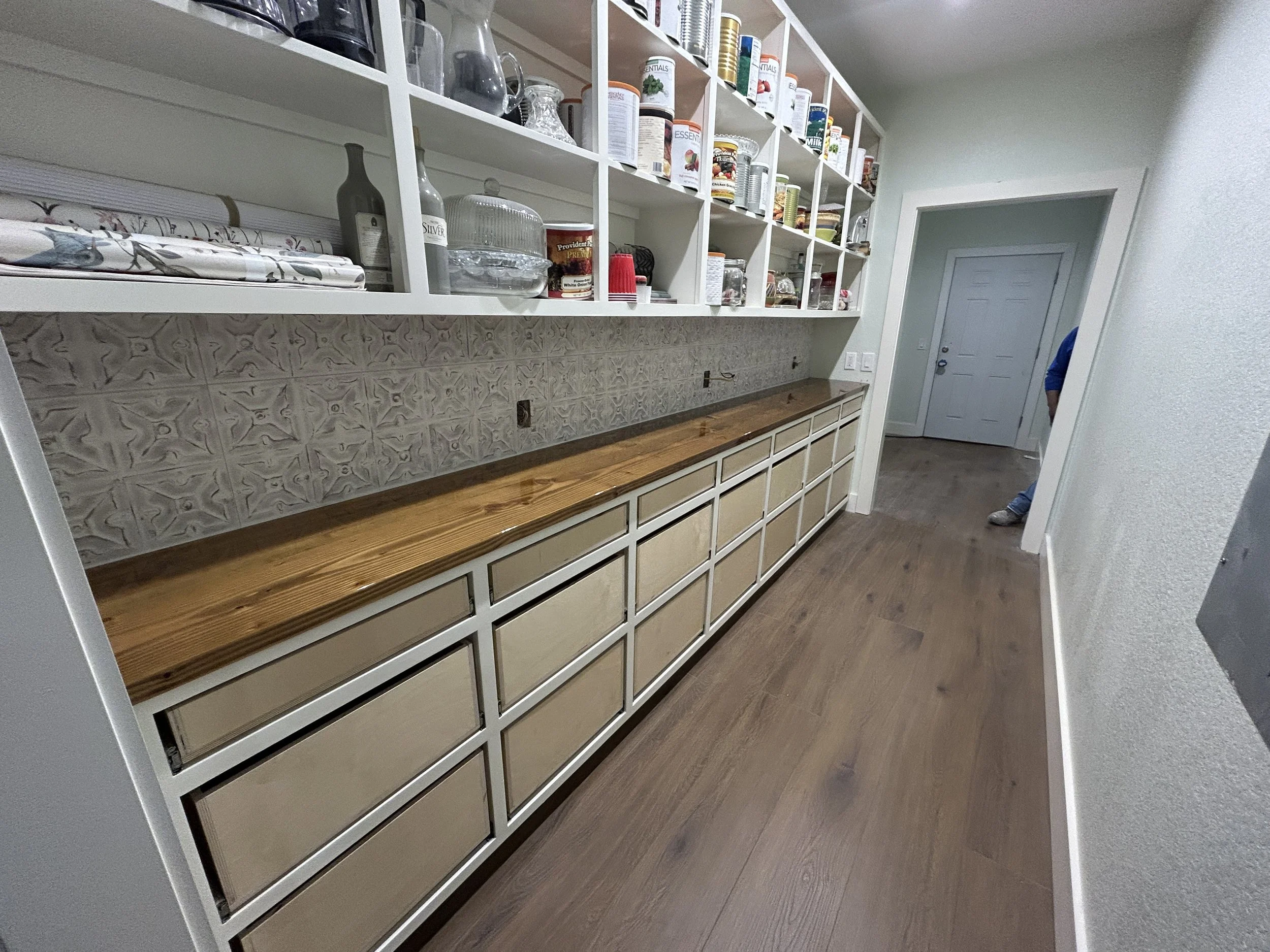 Kitchen pantry with open white shelves filled with jars, cans, and boxes, and a long wooden counter with drawers below, against a decorated backsplash and a white wall.
