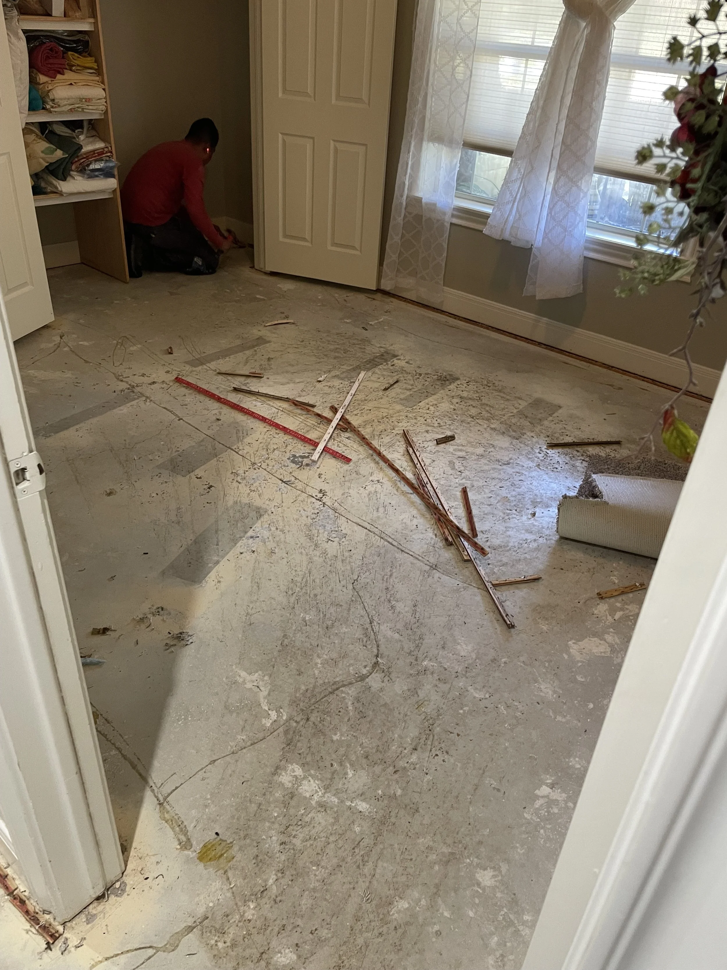 Room with a person working on flooring renovation, scattered wood debris, construction tools, and a window with curtains.