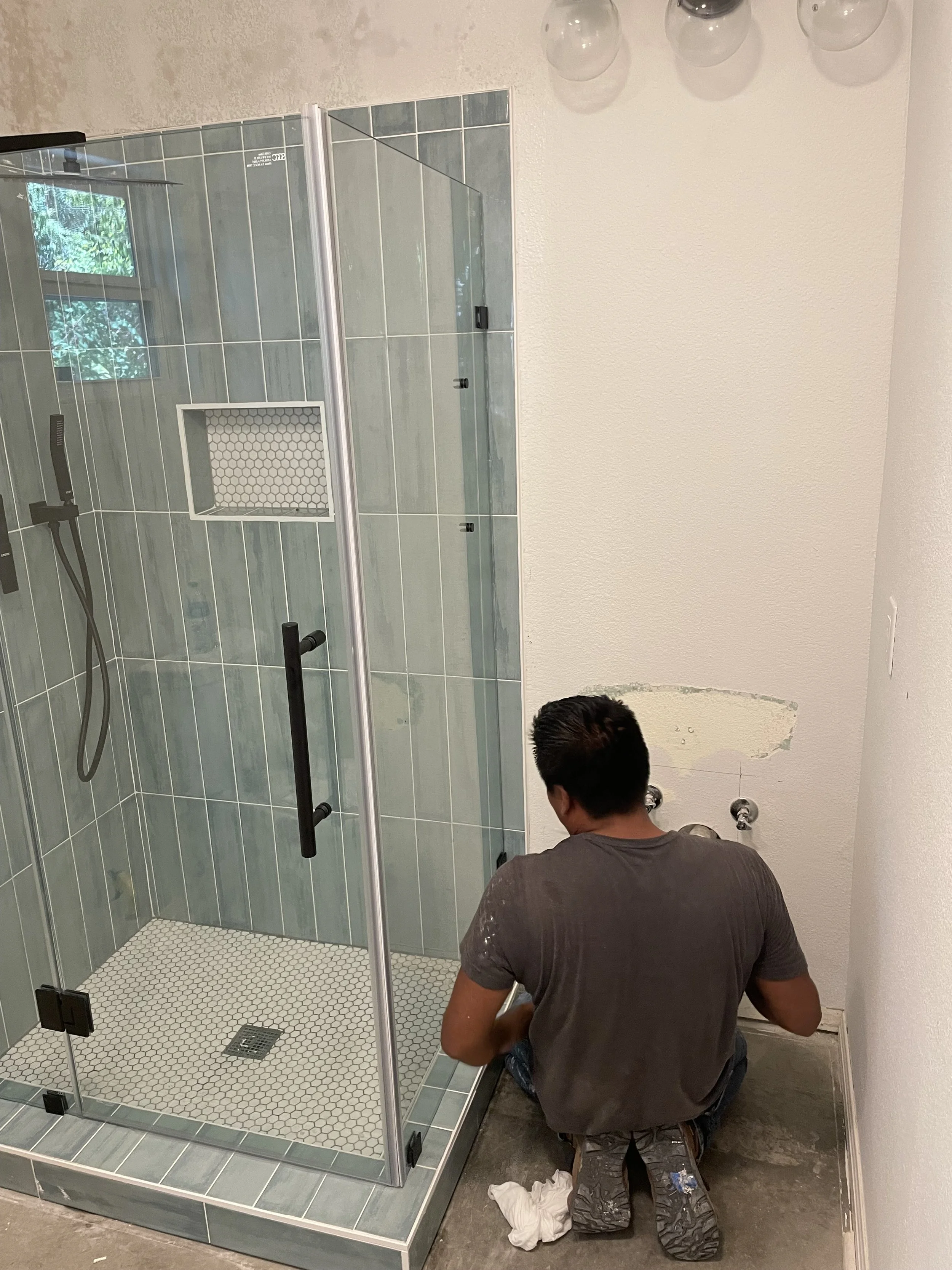 A man working on installing or repairing a bathroom shower with green tiles, a glass door, and a small wall niche, in front of a white wall with some damage.