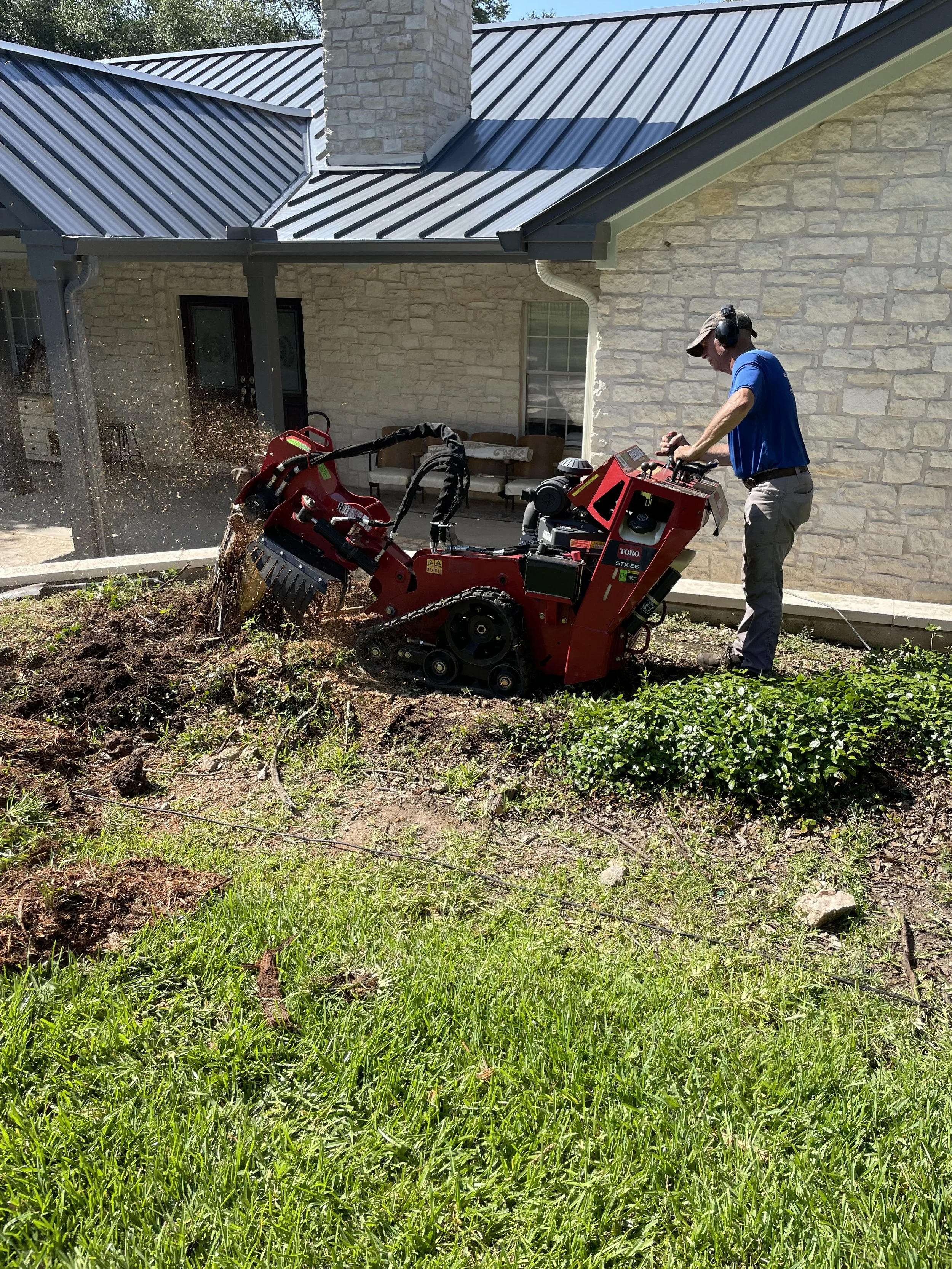 A man operating a red trencher machine on a garden slope in front of a house with stone walls and a metal roof, digging into the soil with dirt and grass around.