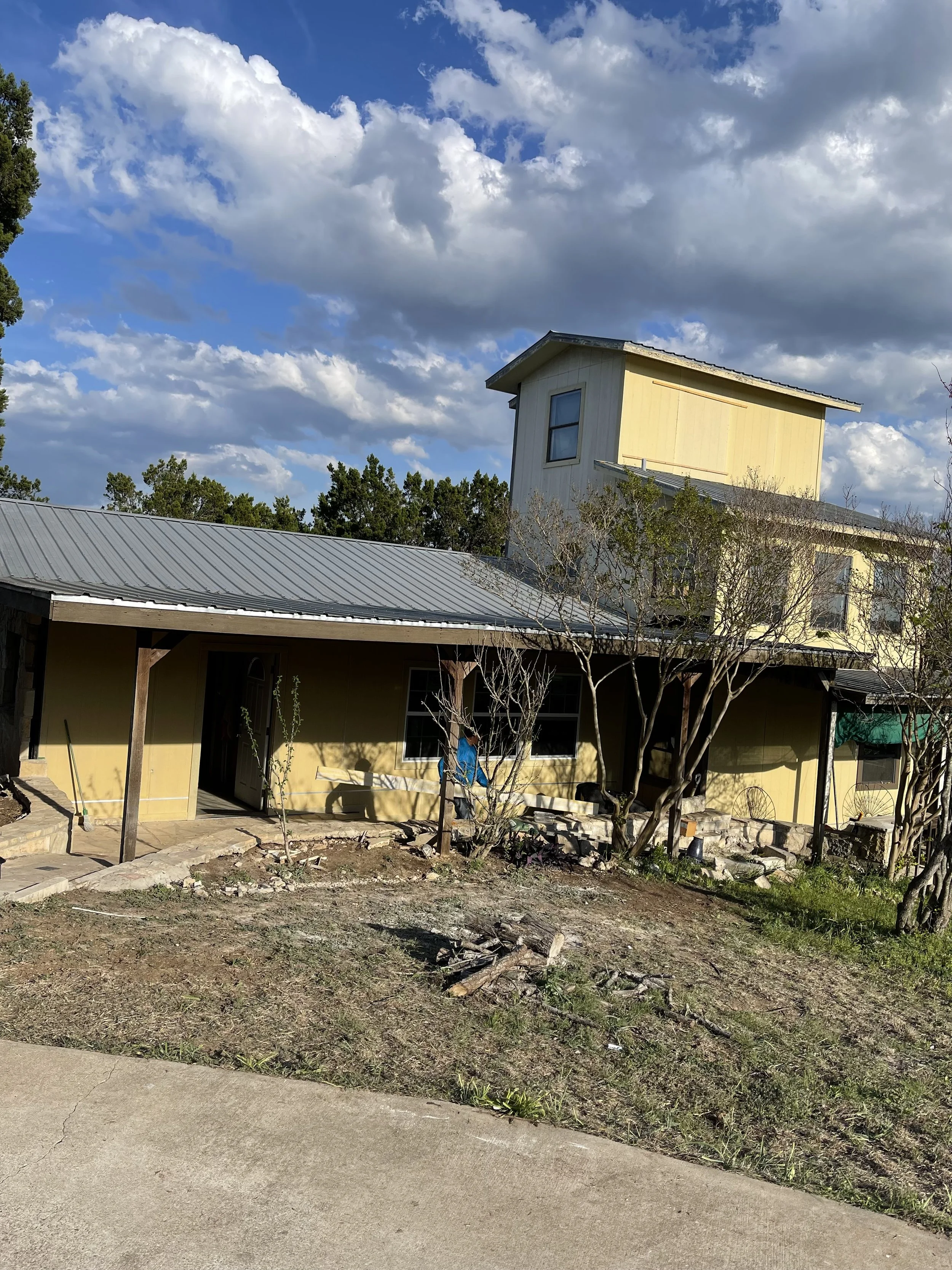 A two-story yellow house with a metal roof, trees in front, and a cloudy sky above.