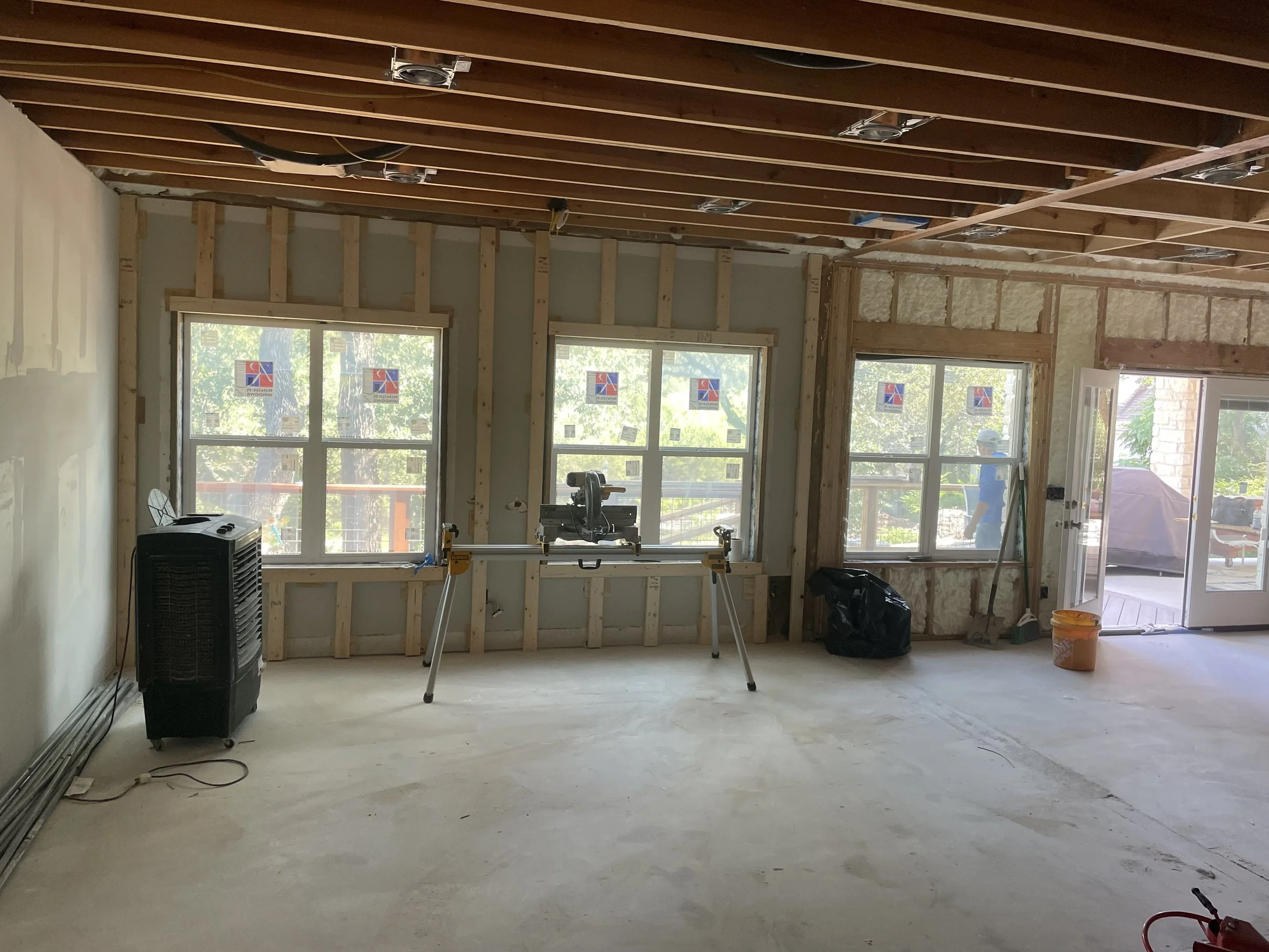 Interior of a room under construction with exposed wooden beams, partially finished walls, windows, and construction tools including a miter saw on a stand.