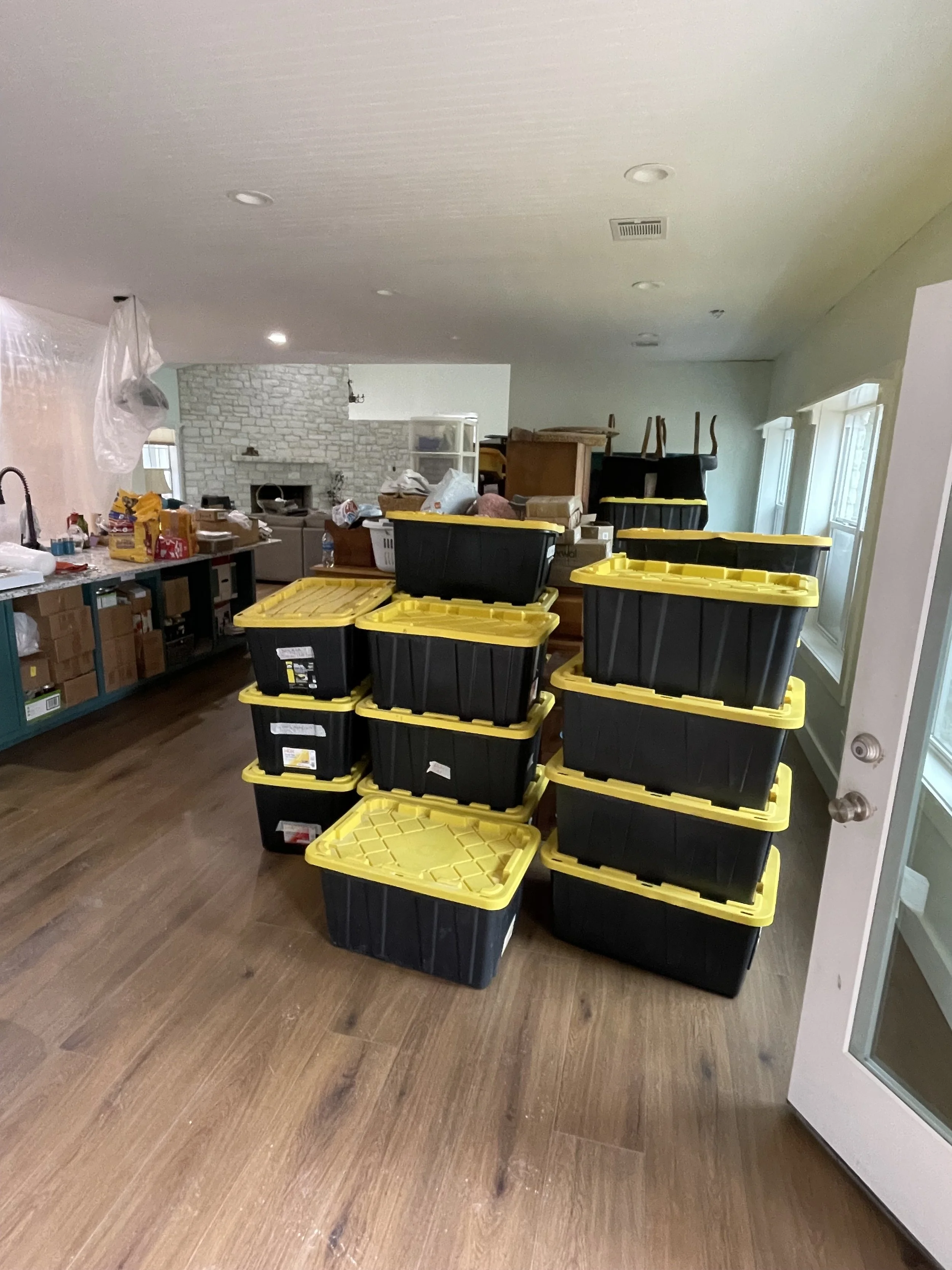 Stacked black and yellow plastic storage bins in a cluttered room with wooden floors and a white brick fireplace in the background.