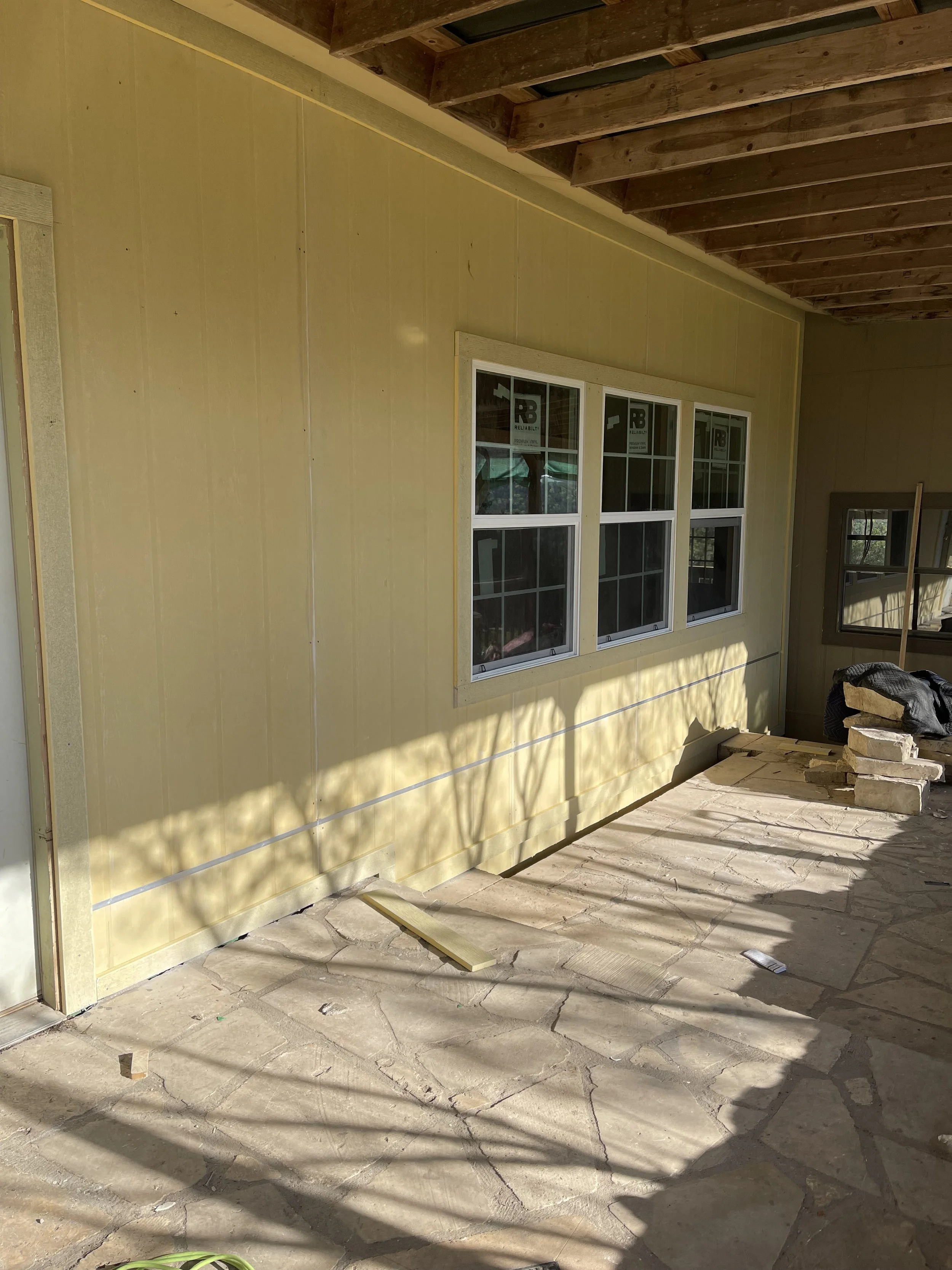 Construction site with a yellow exterior wall, window installed, and unfinished wooden ceiling beams. The floor is covered with large, irregular stone tiles, and shadows from nearby trees are cast on the ground.