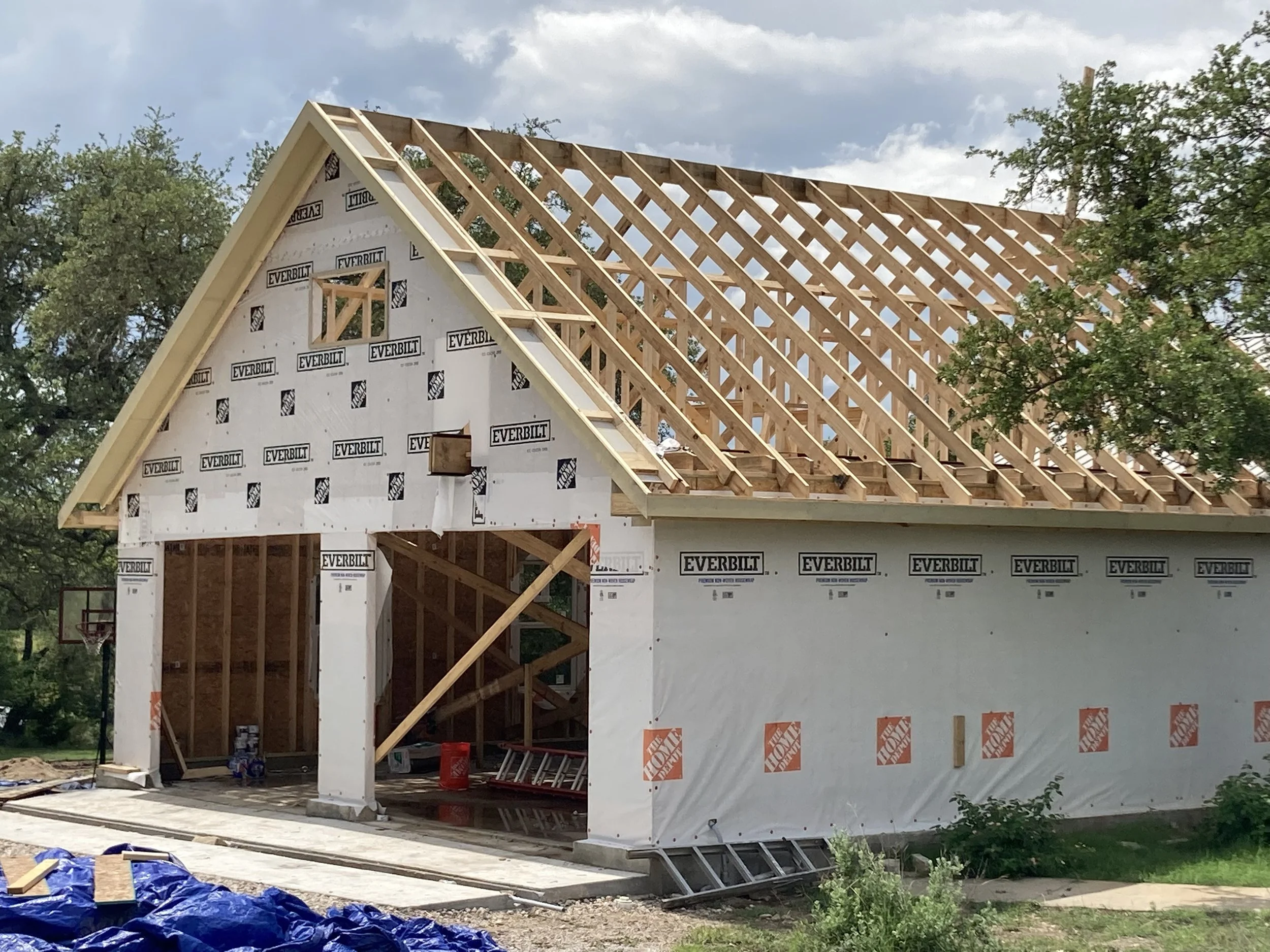 Under-construction house with exposed wooden framing and roof trusses, wrap-around porch opening, and construction materials on site, with trees and sky in background.