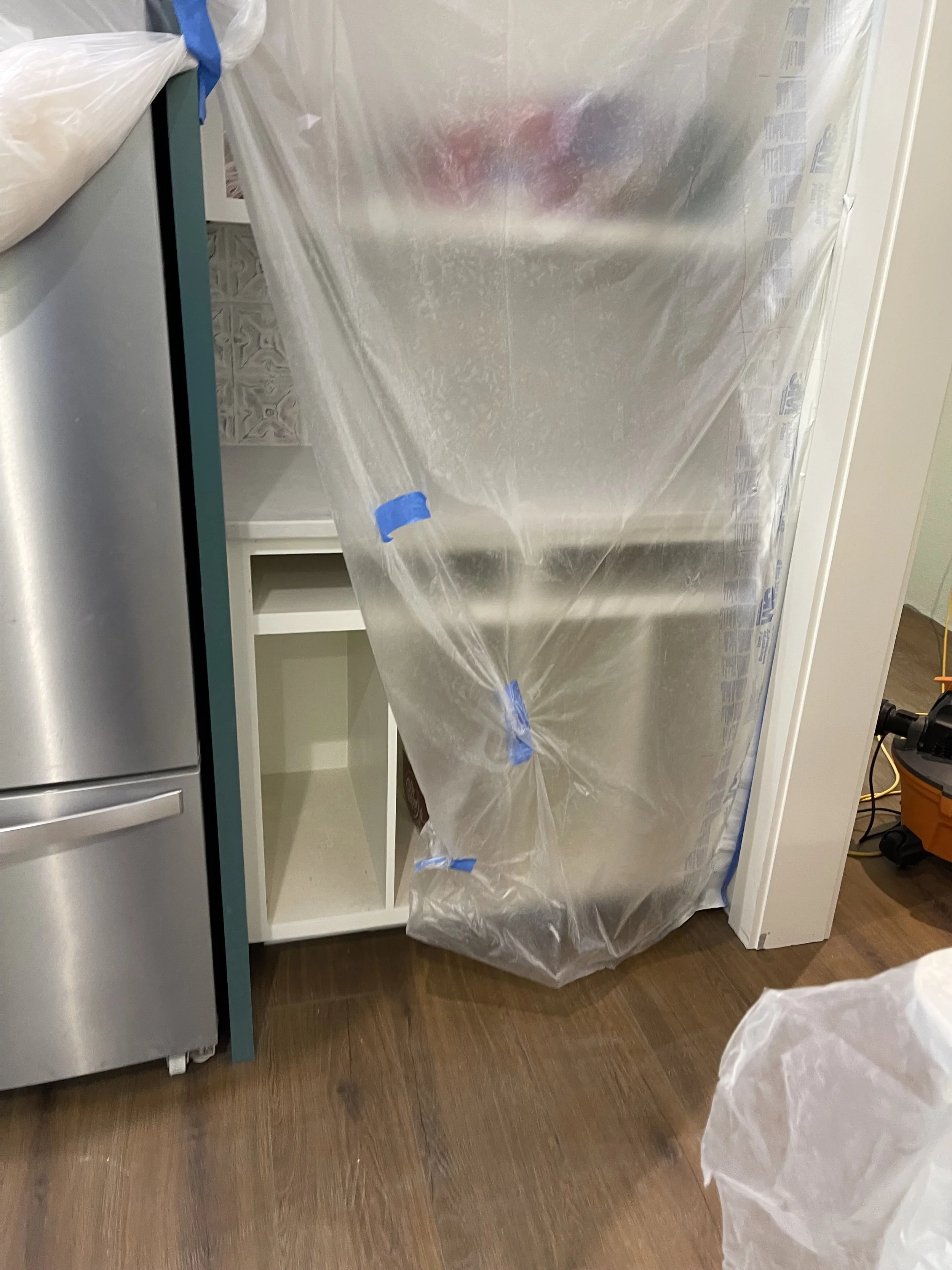 Kitchen with a large plastic sheet hanging in front of a white cabinet and a gray refrigerator, taped at the top and bottom.
