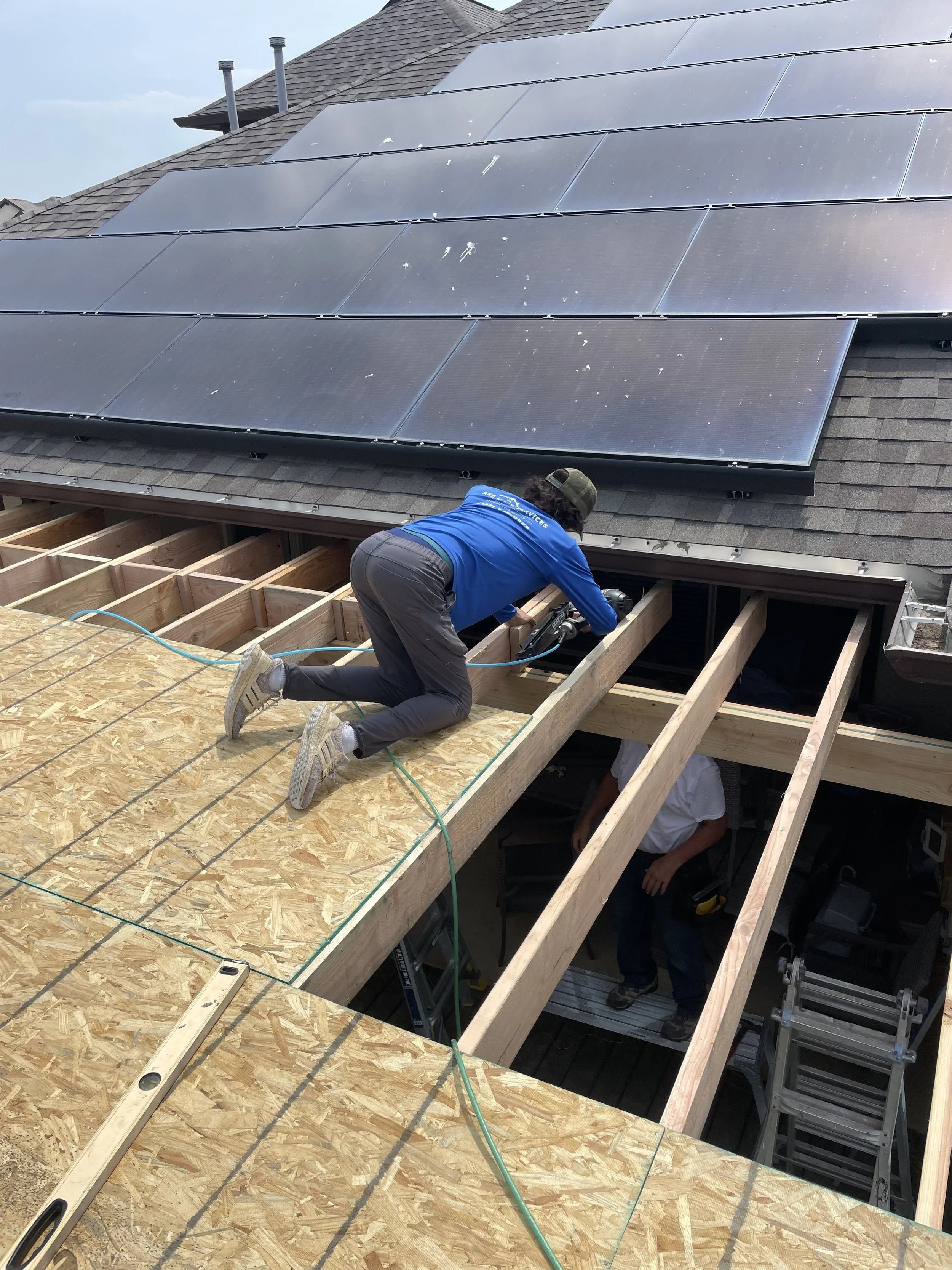 A construction worker in a blue shirt and dark pants installing or repairing wooden beams on a house roof. The roof has solar panels and the worker is kneeling on a wooden platform, working with tools. Another worker is partially visible below, stand
