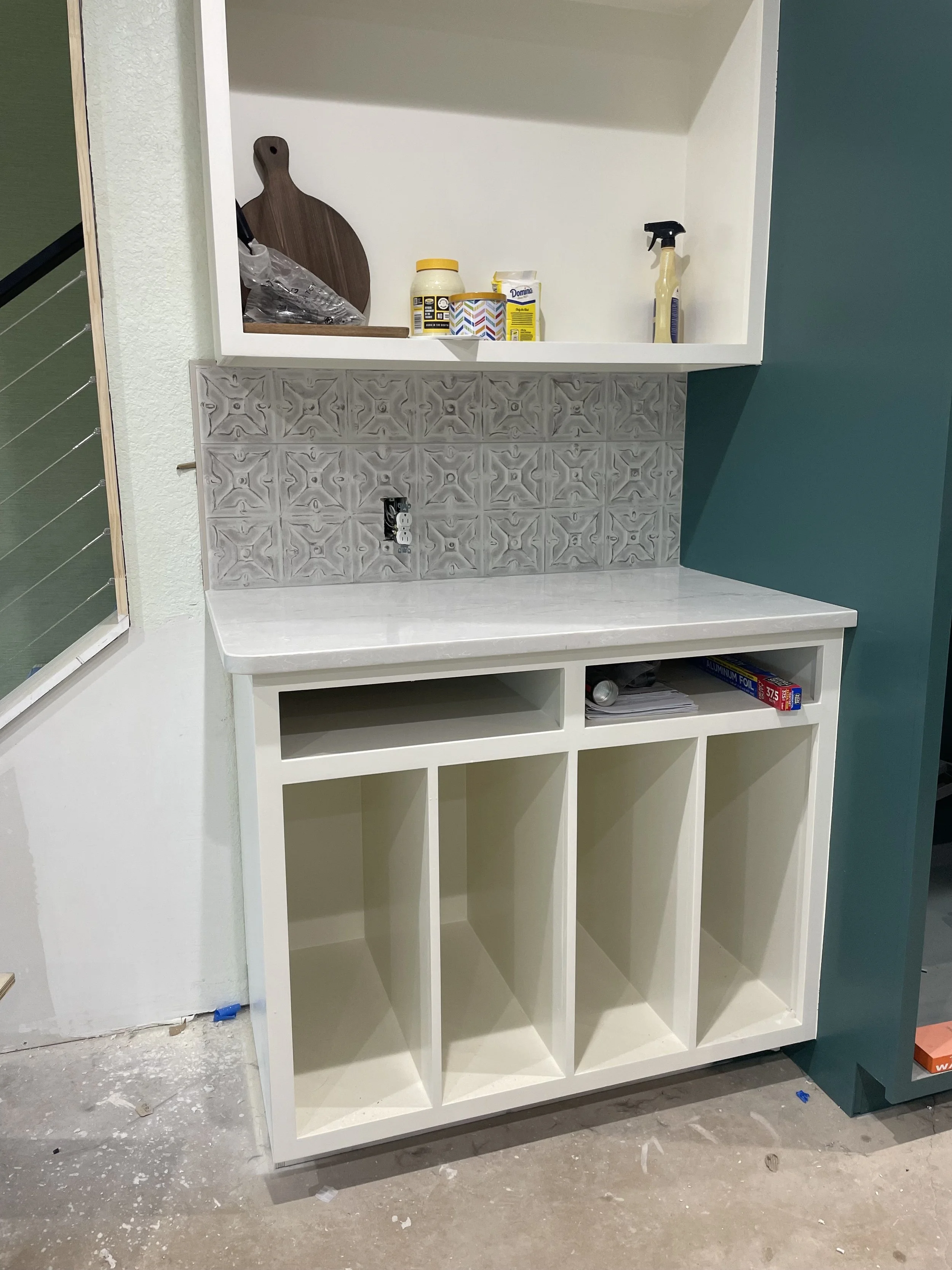 Empty kitchen cabinet with open shelves and a counter, partially tiled wall, and various items on the upper shelf, including a cutting board, condiment bottle, spray bottle, and cleaning supplies.