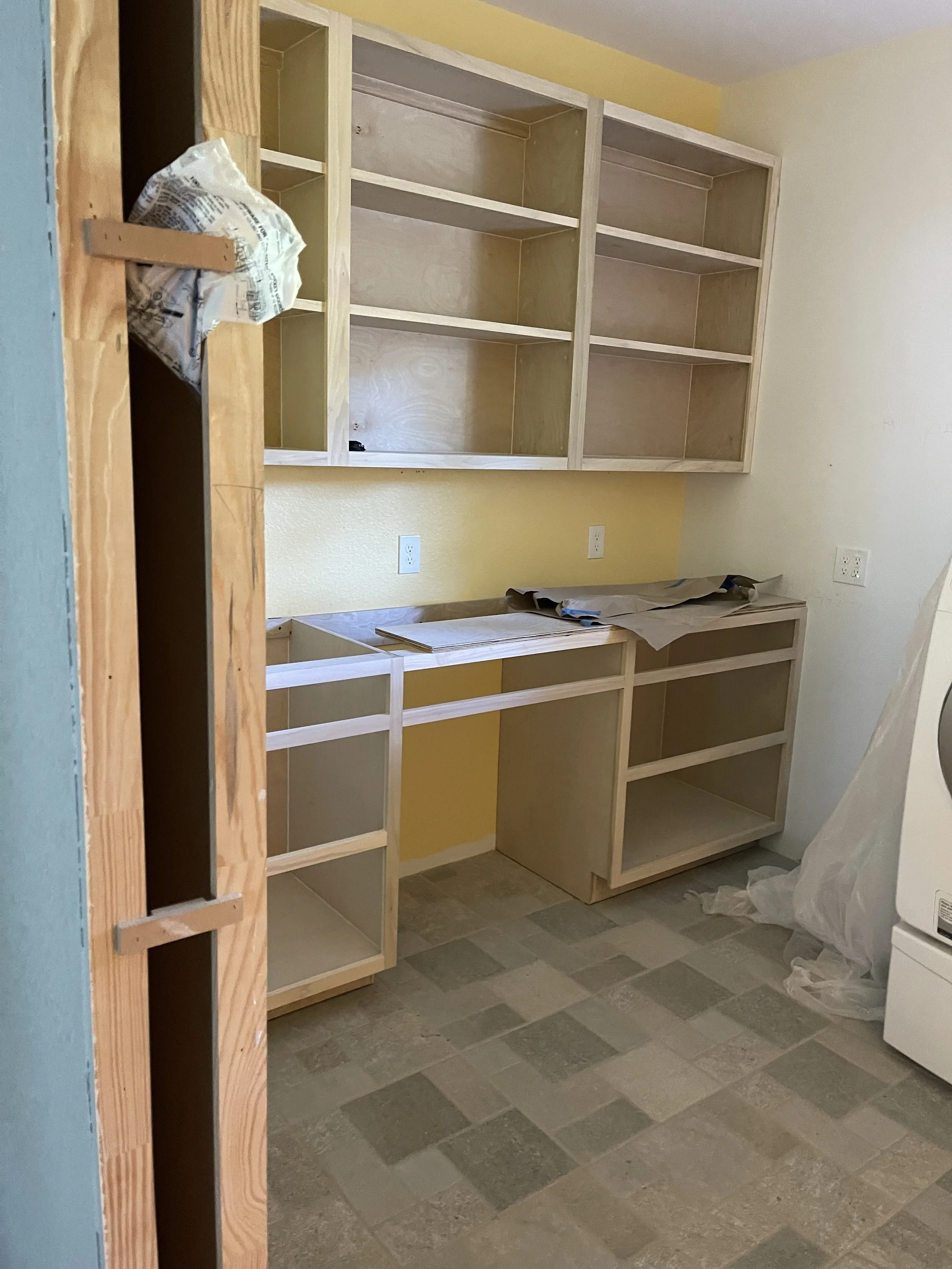 Unfinished kitchen or utility room with wood cabinets and an empty countertop, yellow painted wall, electrical outlets, and a washing machine partially covered with plastic.