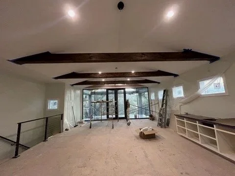 Renovated open-concept room with exposed wood ceiling beams, large glass doors, recessed lighting, ladder, and built-in shelving.