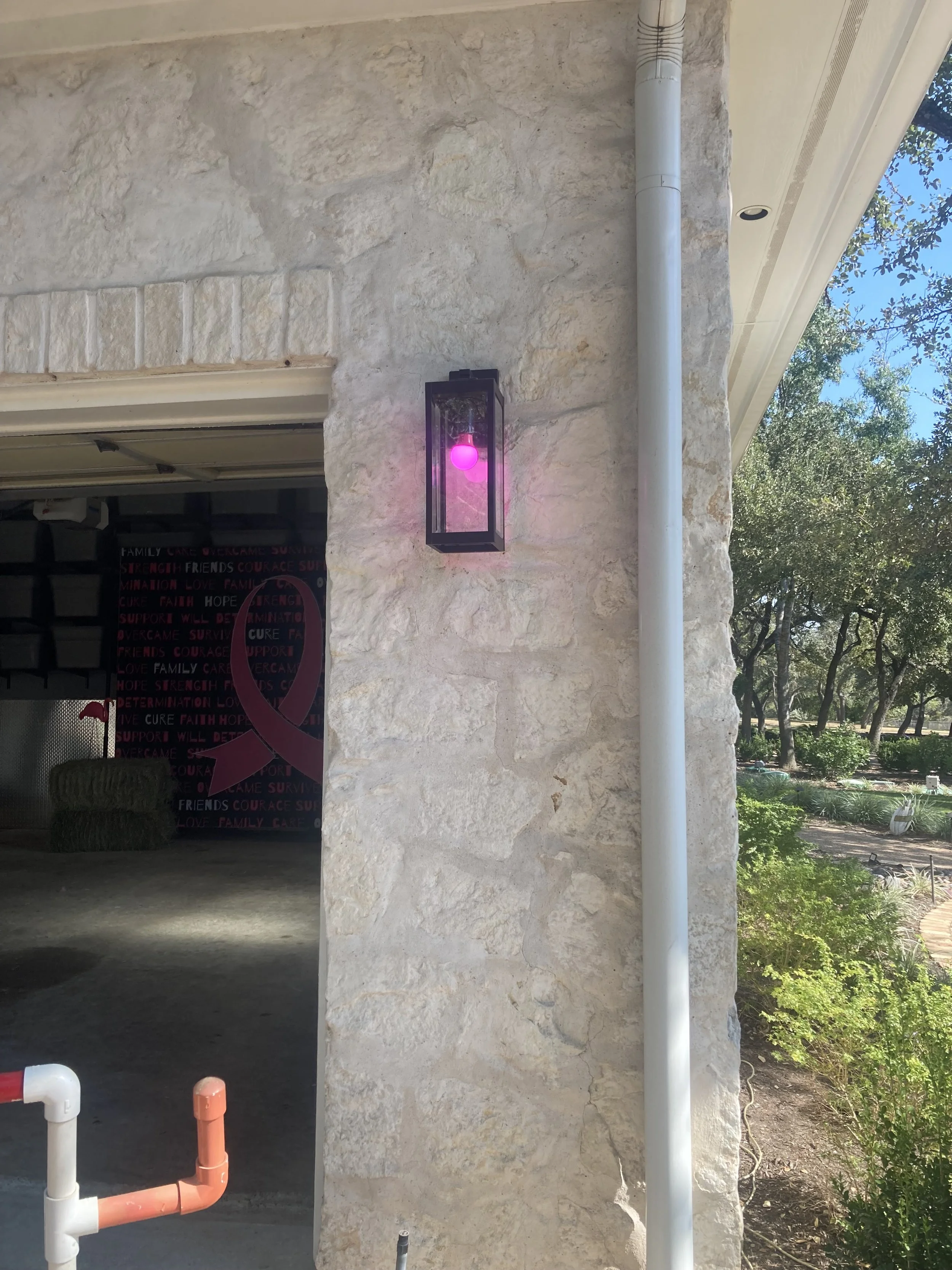 Exterior stone wall with a black lantern-style light fixture emitting a pink glow, a white downspout, and a partly visible red and white pipe. The background shows part of a garden with trees and blue sky.