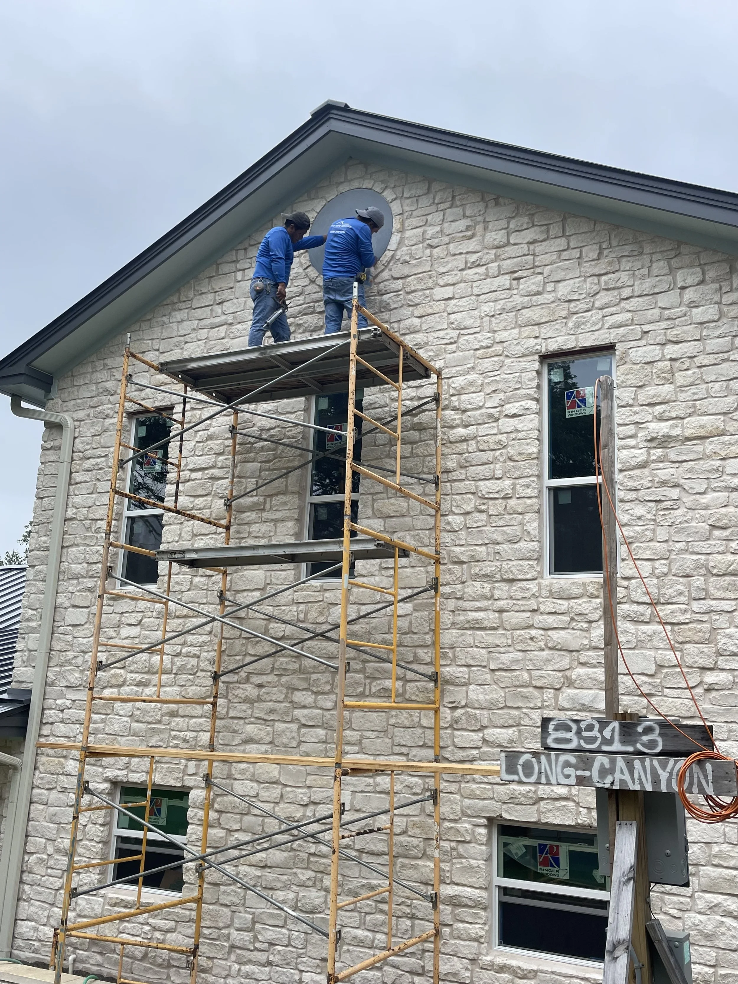 Two workers on scaffolding installing or repairing a circular window near the roof of a stone house.
