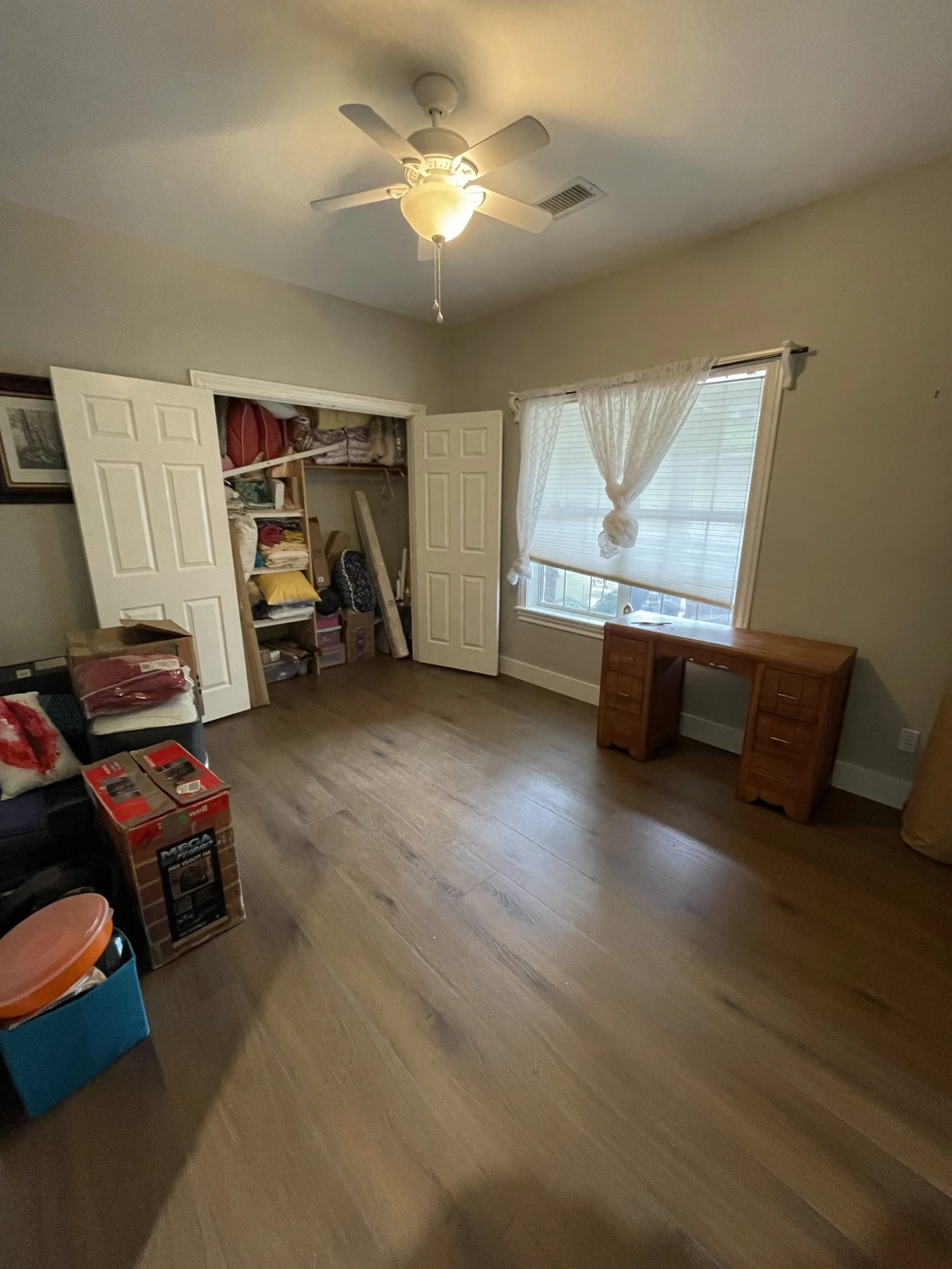 Empty cluttered bedroom with wooden floor, open closet filled with items, a desk near the window with sheer curtains, a ceiling fan with light, and some clutter on the floor.