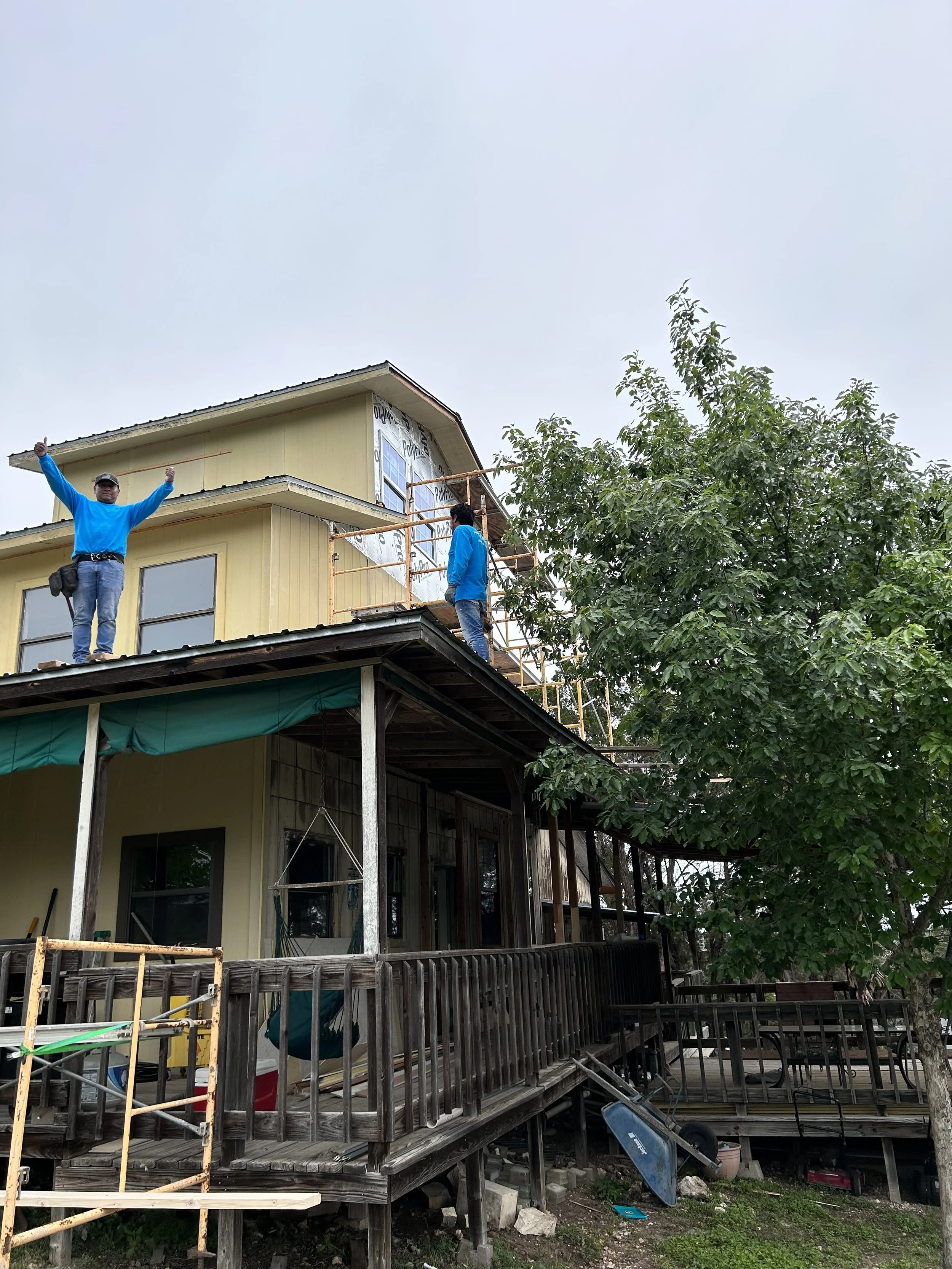Two workers on a house's roof, one standing on the edge with arms raised, the other working with scaffolding around the upper part, surrounded by trees and a wooden porch.
