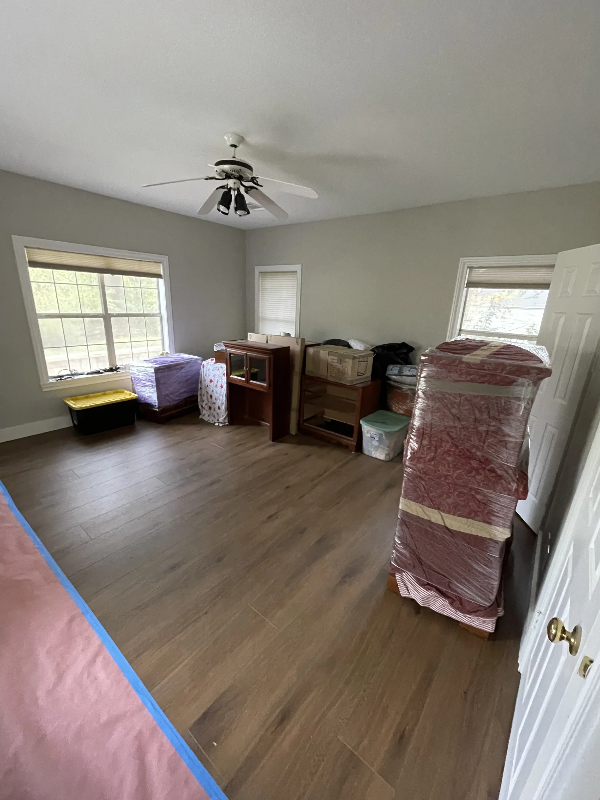 A room with hardwood floors, three windows with blinds, and a ceiling fan. The room contains various furniture and boxes, some covered in plastic wrap.