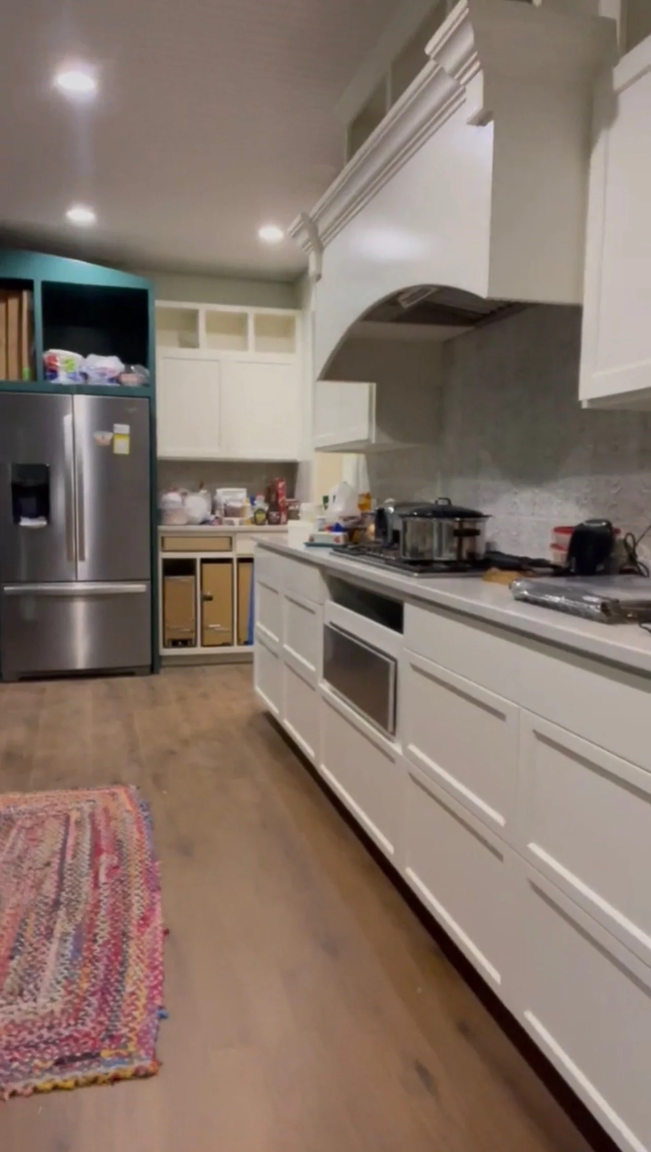 A kitchen with white cabinets, a stainless steel refrigerator, and a wooden floor. There is a colorful rug on the floor, and the countertops have various kitchen appliances and items.