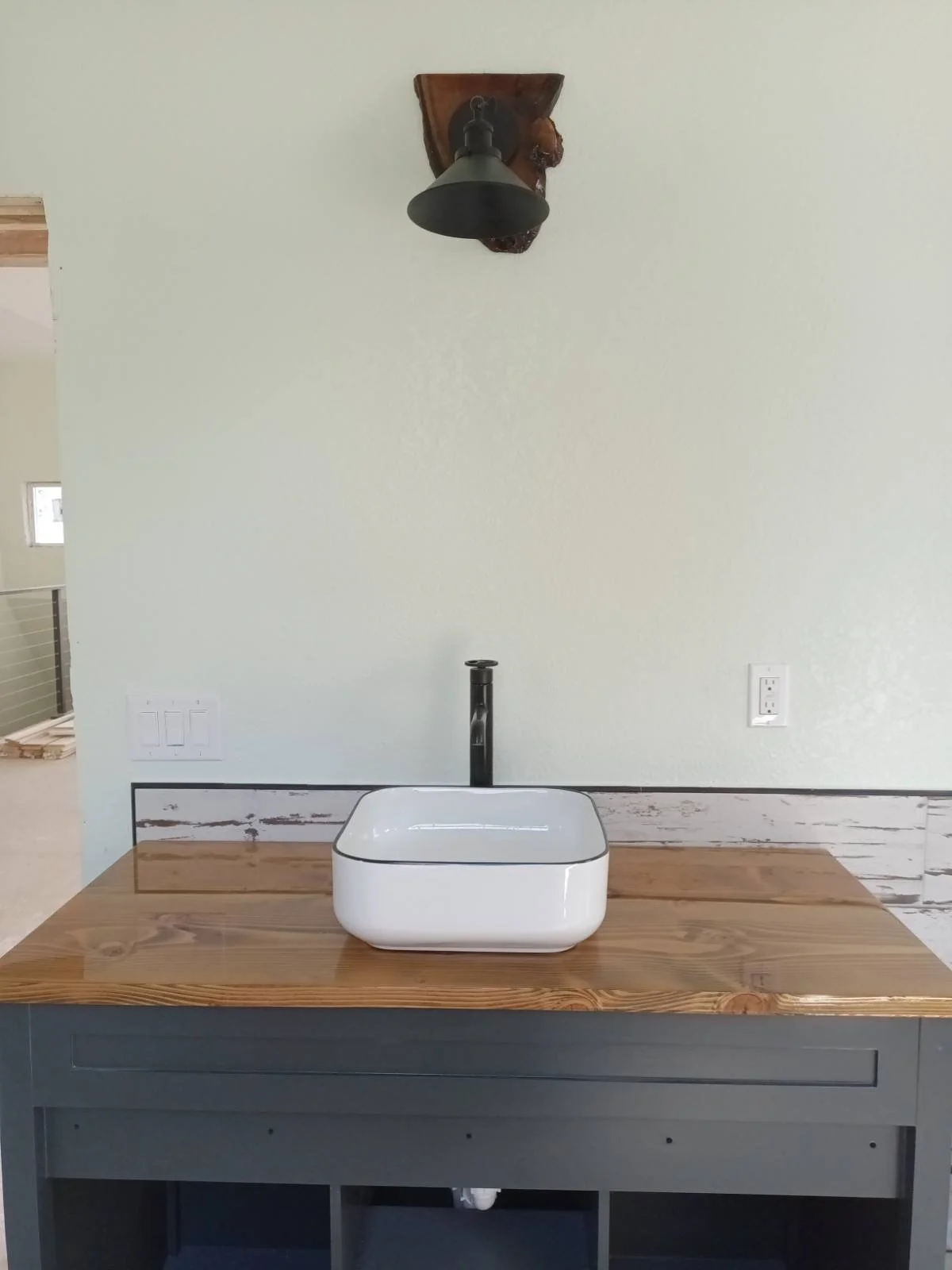 Bathroom vanity with a white rectangular vessel sink on a wooden countertop, black faucet in the center, and a black wall-mounted lamp overhead, with a light green wall and electrical outlets behind.