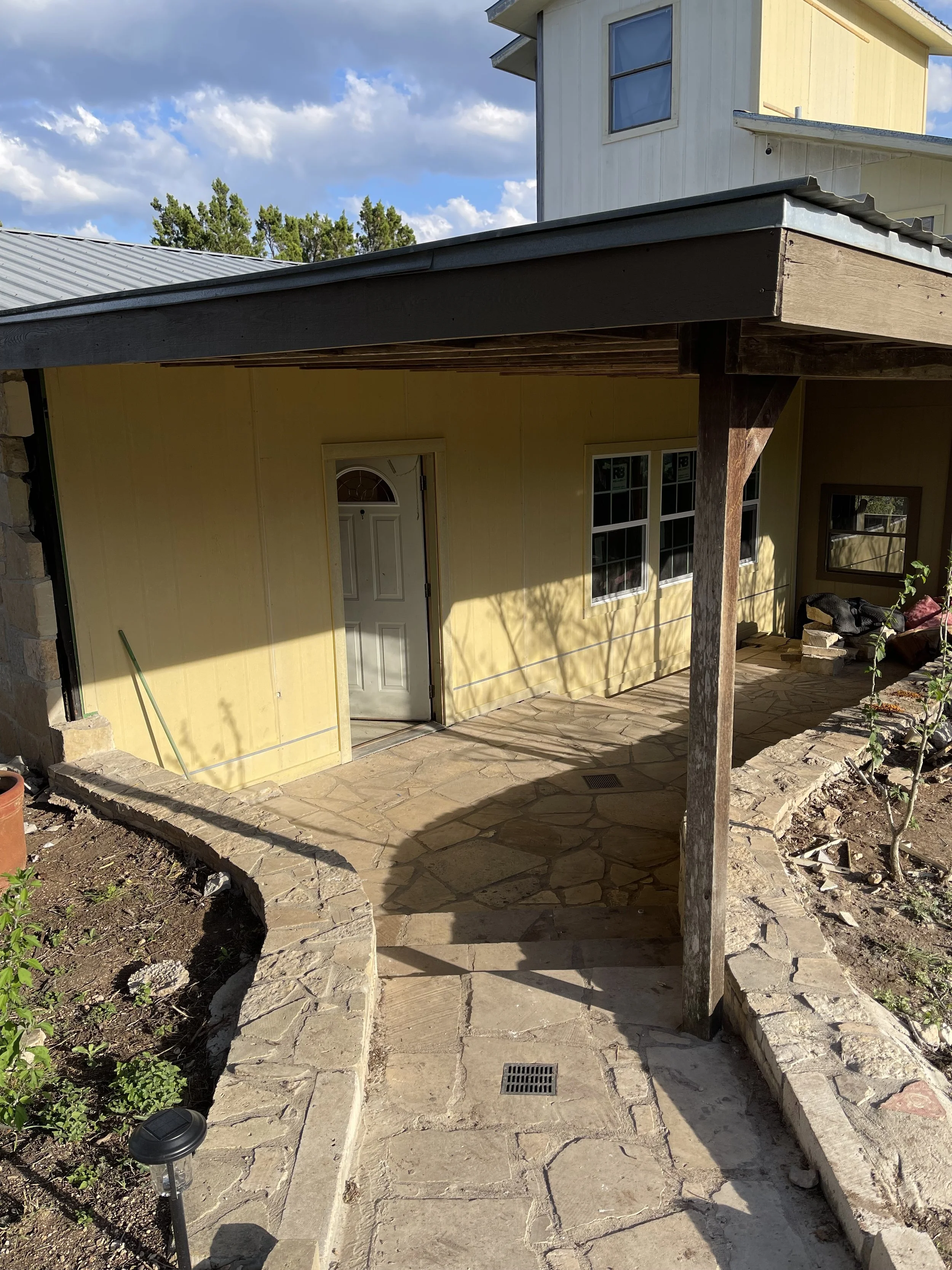 Backyard patio with stone walkway leading to a yellow house with a white door and black window frames, partially shaded by a roof extension, with some gardening tools and furniture visible.