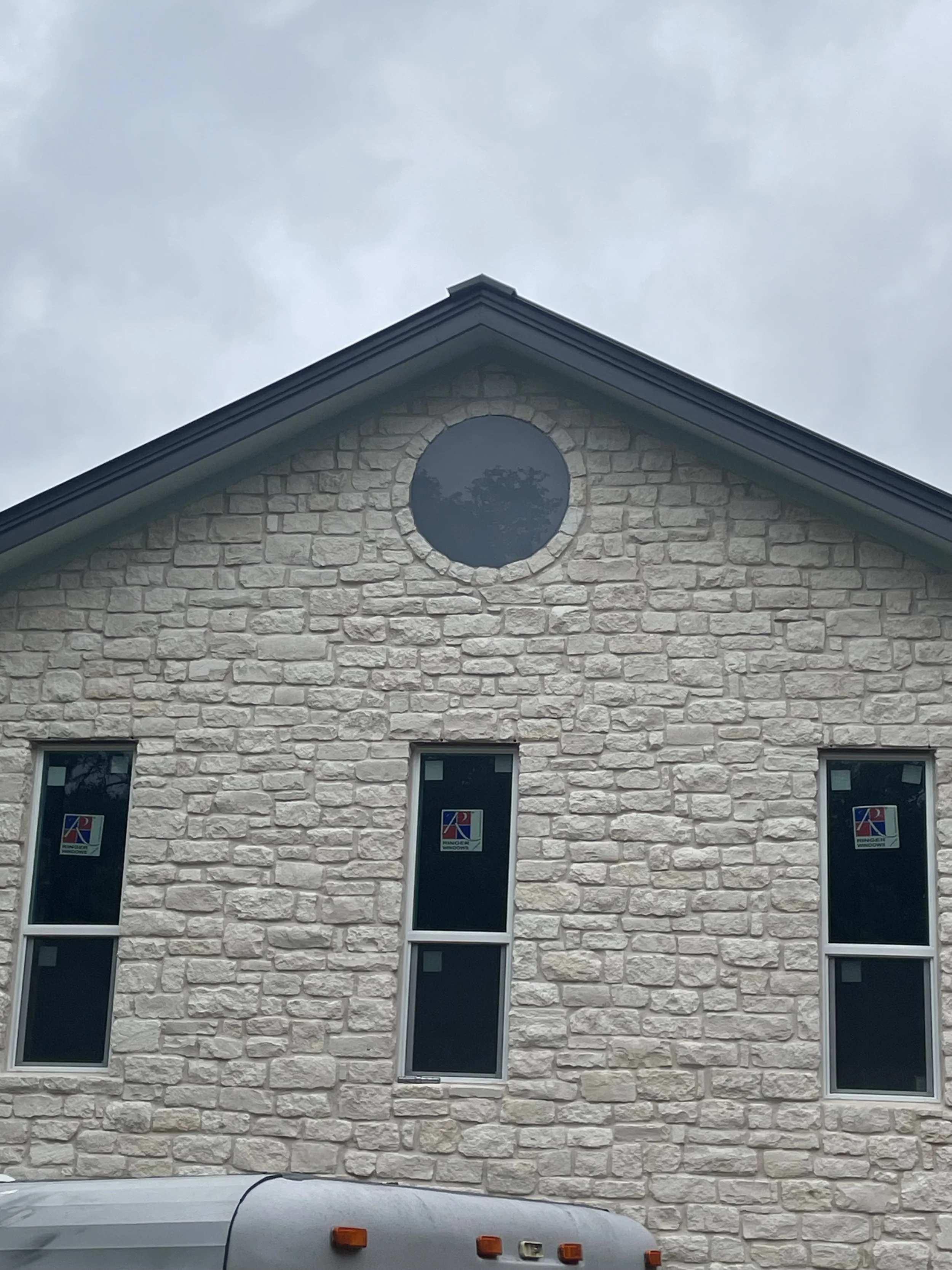 Front view of a stone house with three vertical rectangular windows and a circular window above them, with cloudy sky in background.