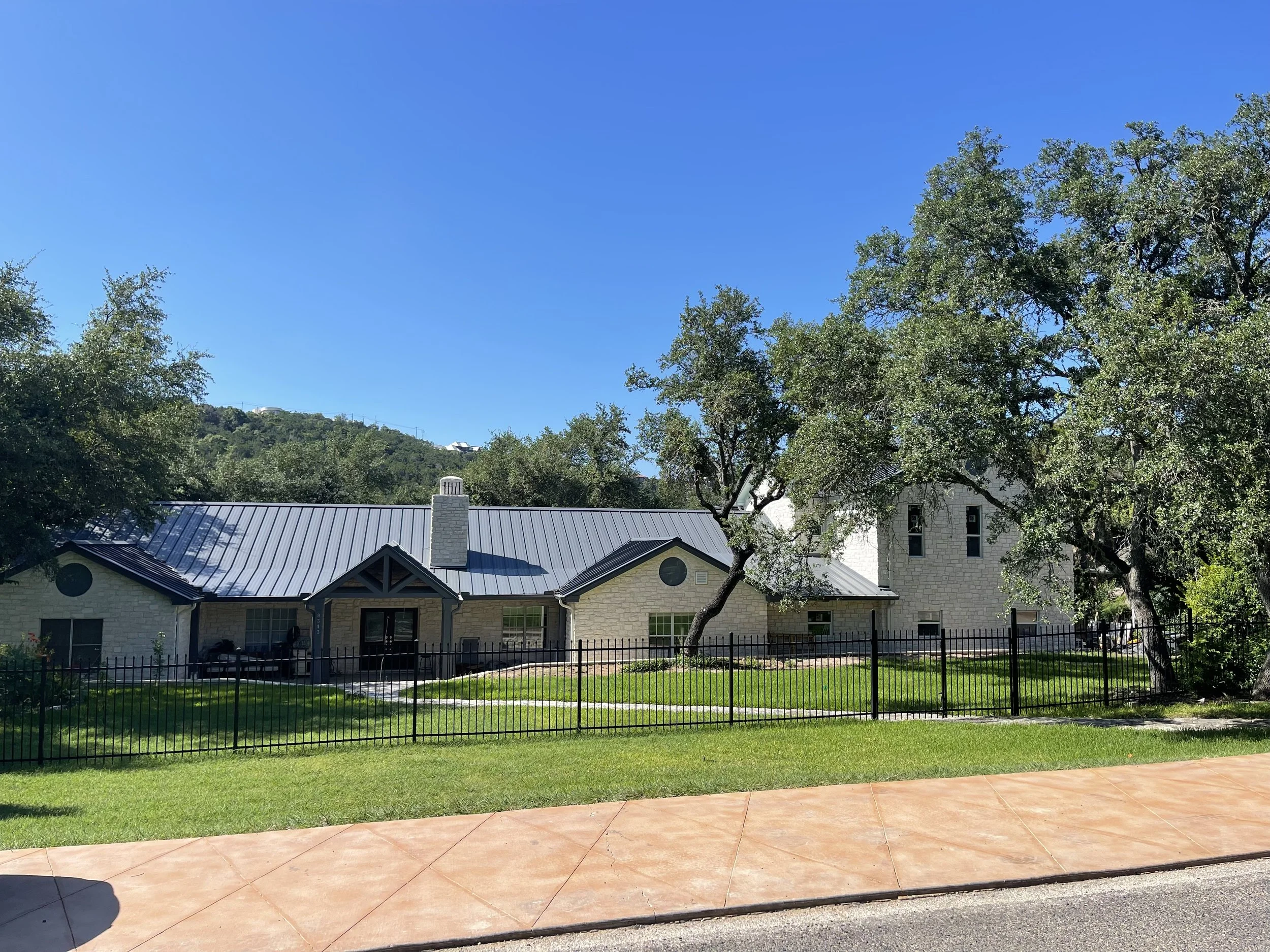 A house with a light-colored brick exterior, a metal roof, and a tall, narrow tower section, set in a landscaped yard with trees and a black metal fence, under a clear blue sky.