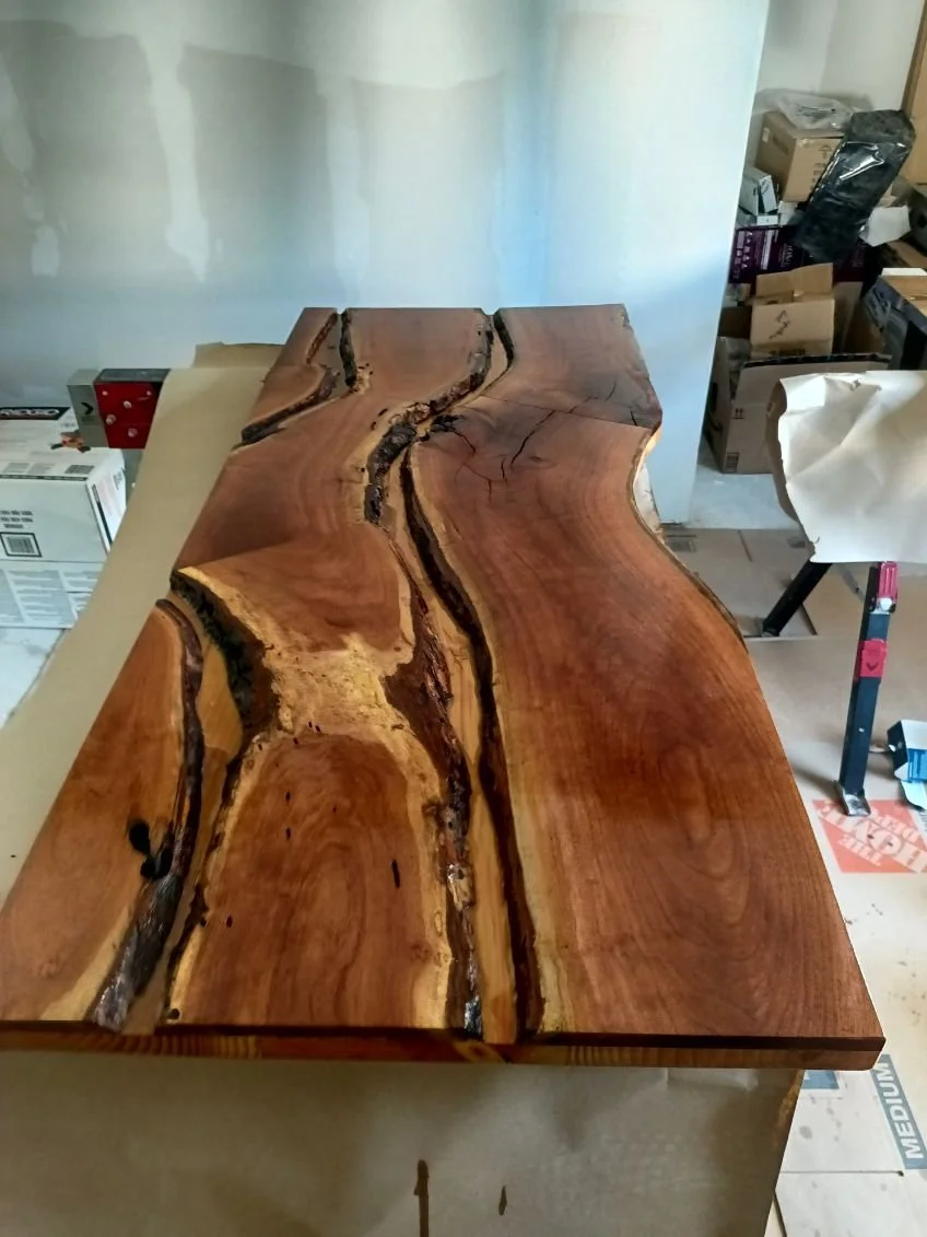 A large slab of natural wood with live edges, showing rich grain patterns and cracks, resting on a worktable in a workshop.