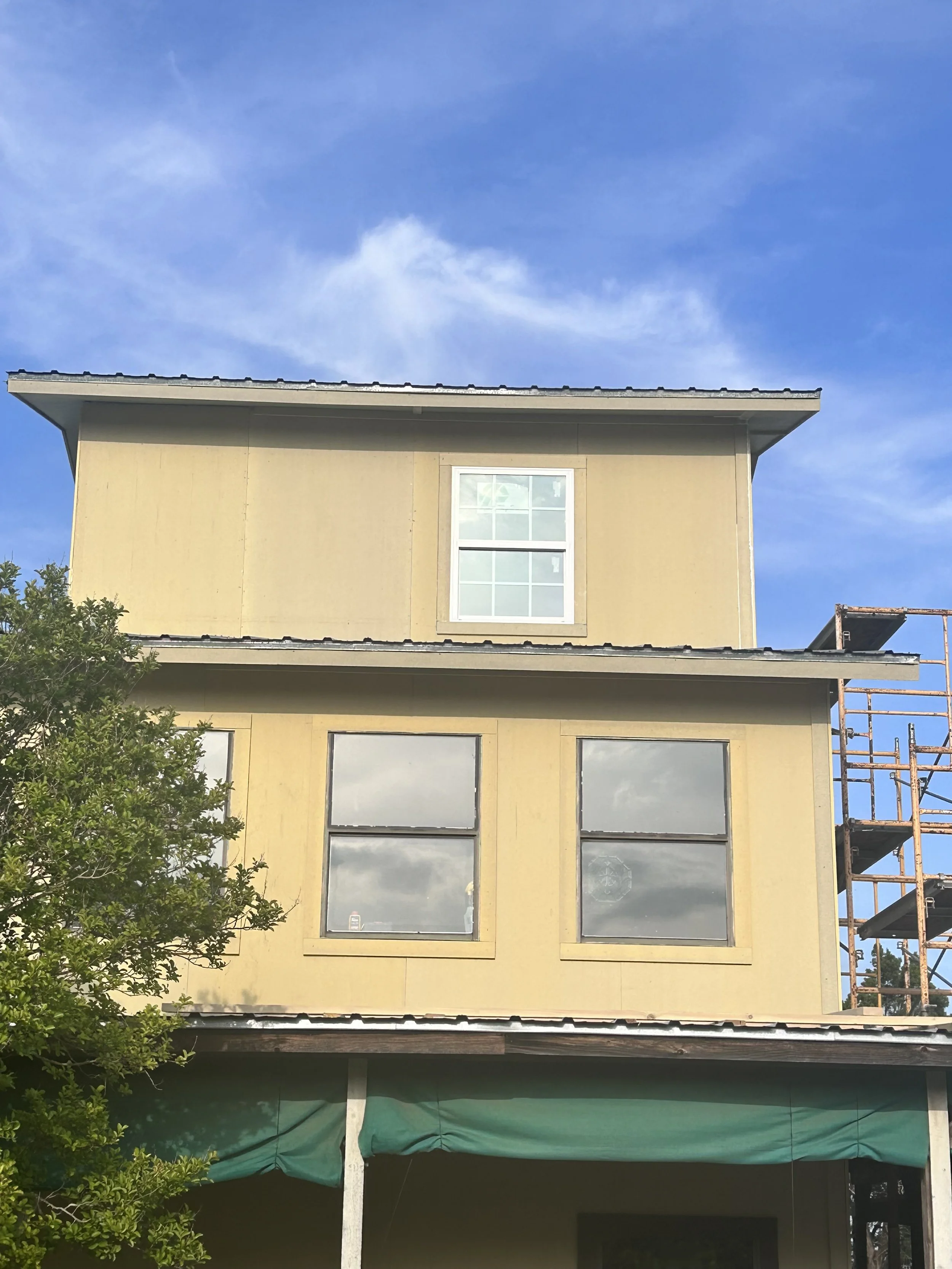 A three-story house under construction with a beige exterior, two windows on the second floor and one window on the third floor, and a scaffold on the right side. The house has a green canopy on the lower level, and a tree is visible on the left side