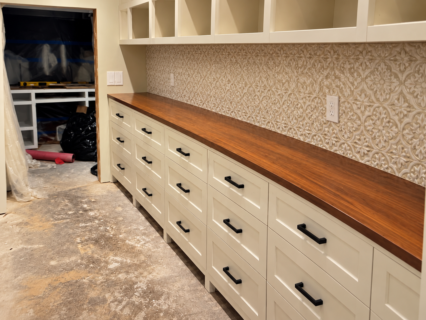Custom cream-colored built-in cabinetry with black hardware, walnut countertop, and ornate patterned tile backsplash in a remodeled room