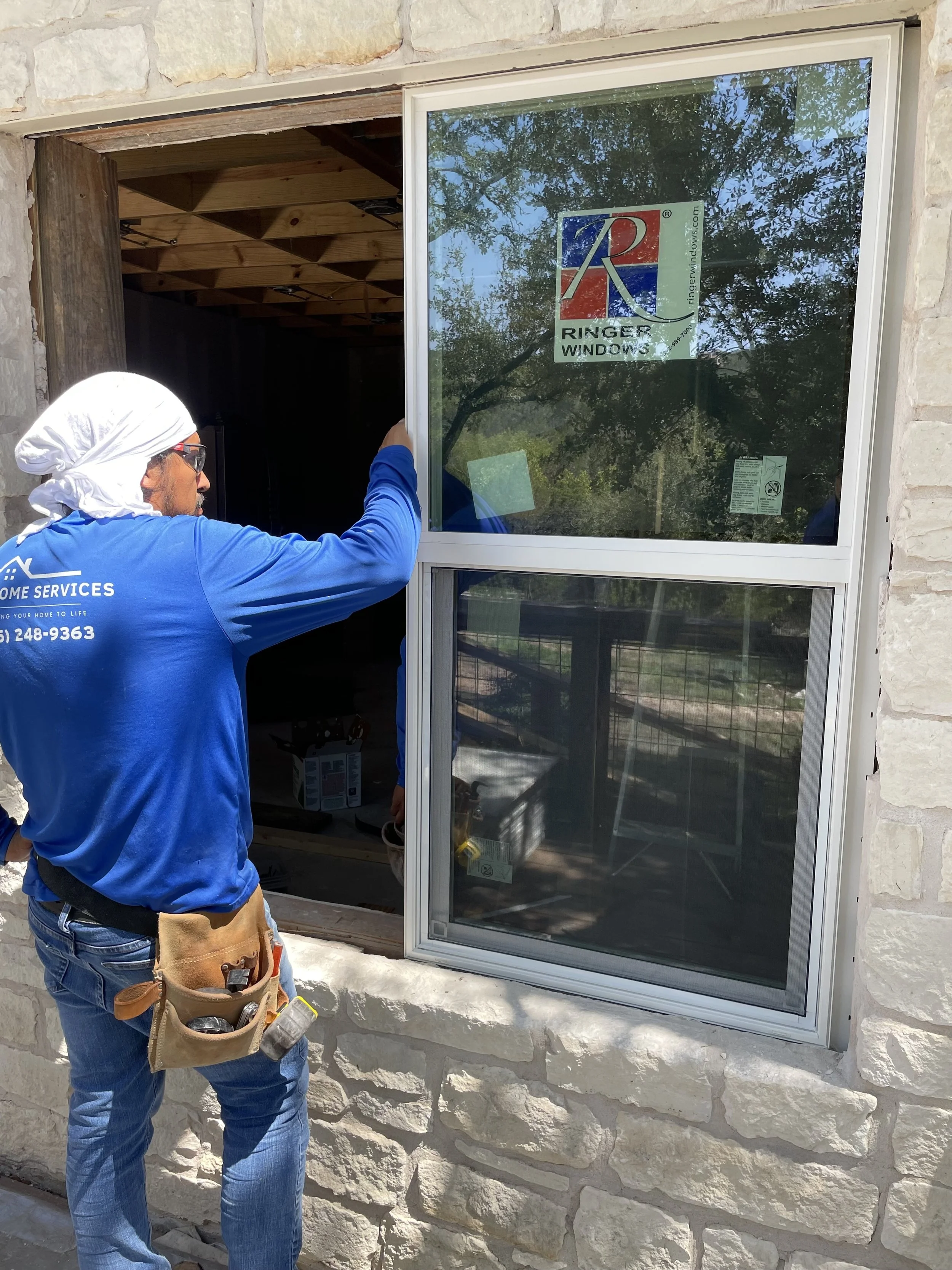A worker installing or repairing a window frame on a stone building exterior. The worker is wearing a blue shirt, jeans, a white head covering, sunglasses, and a tool belt.
