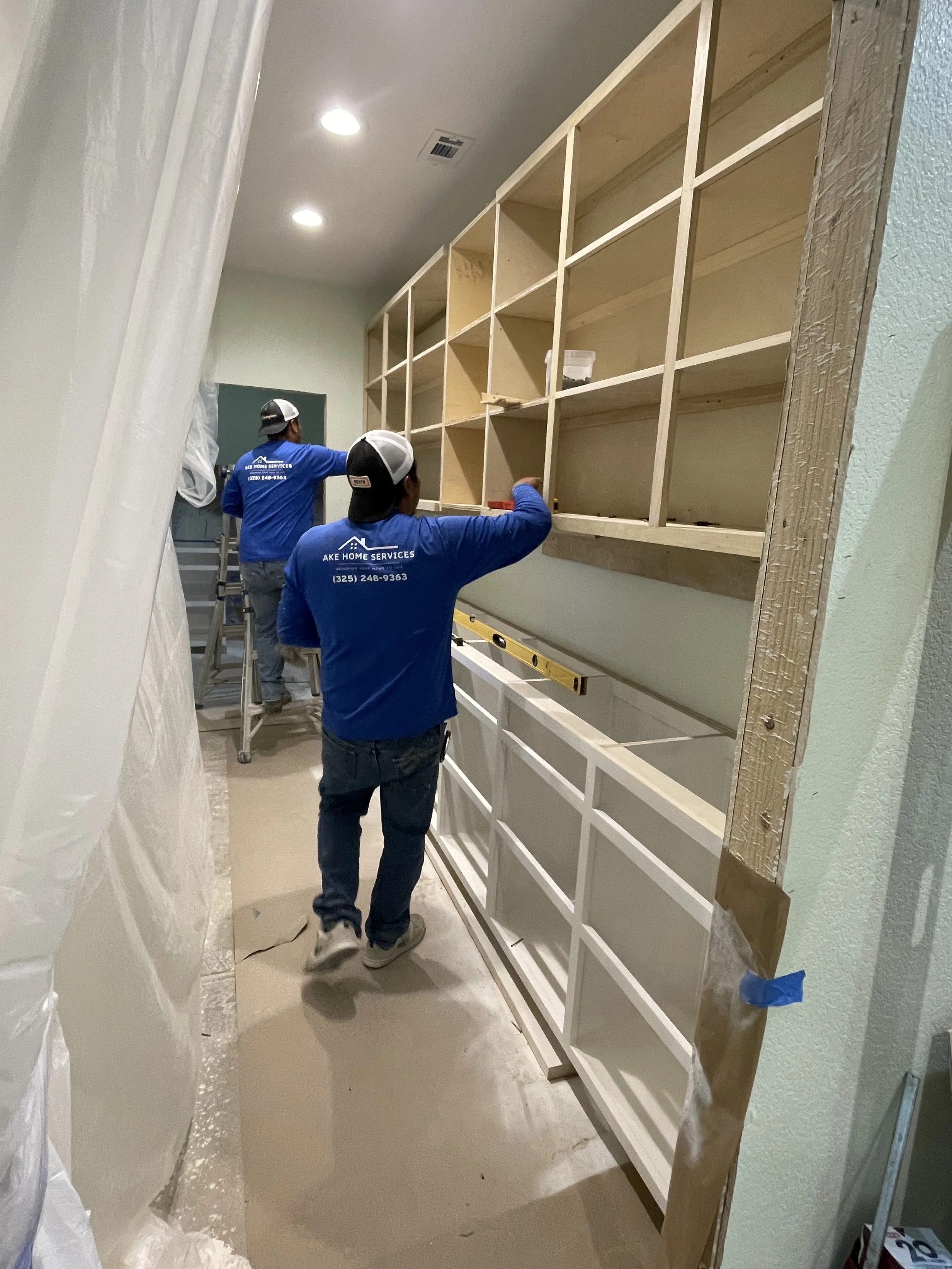 Two workers in blue shirts are installing shelving units in a room, with one on a ladder and the other on the floor. The shelving is unfinished wood, and the workers are working on mounting it to the wall.