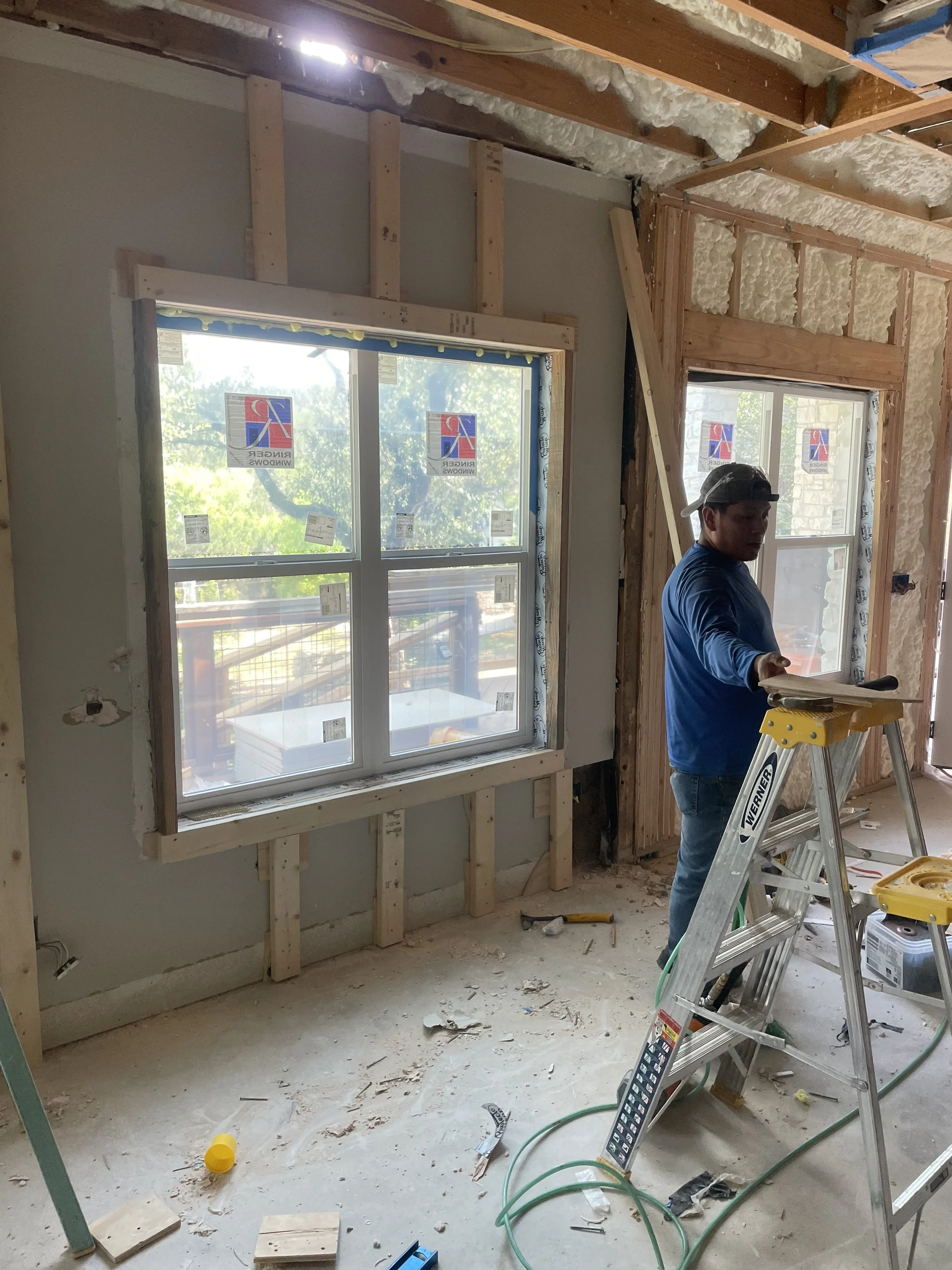 Construction worker inside a house under renovation, installing or inspecting windows, with unfinished walls and ceiling, tools, and building materials scattered on the floor.