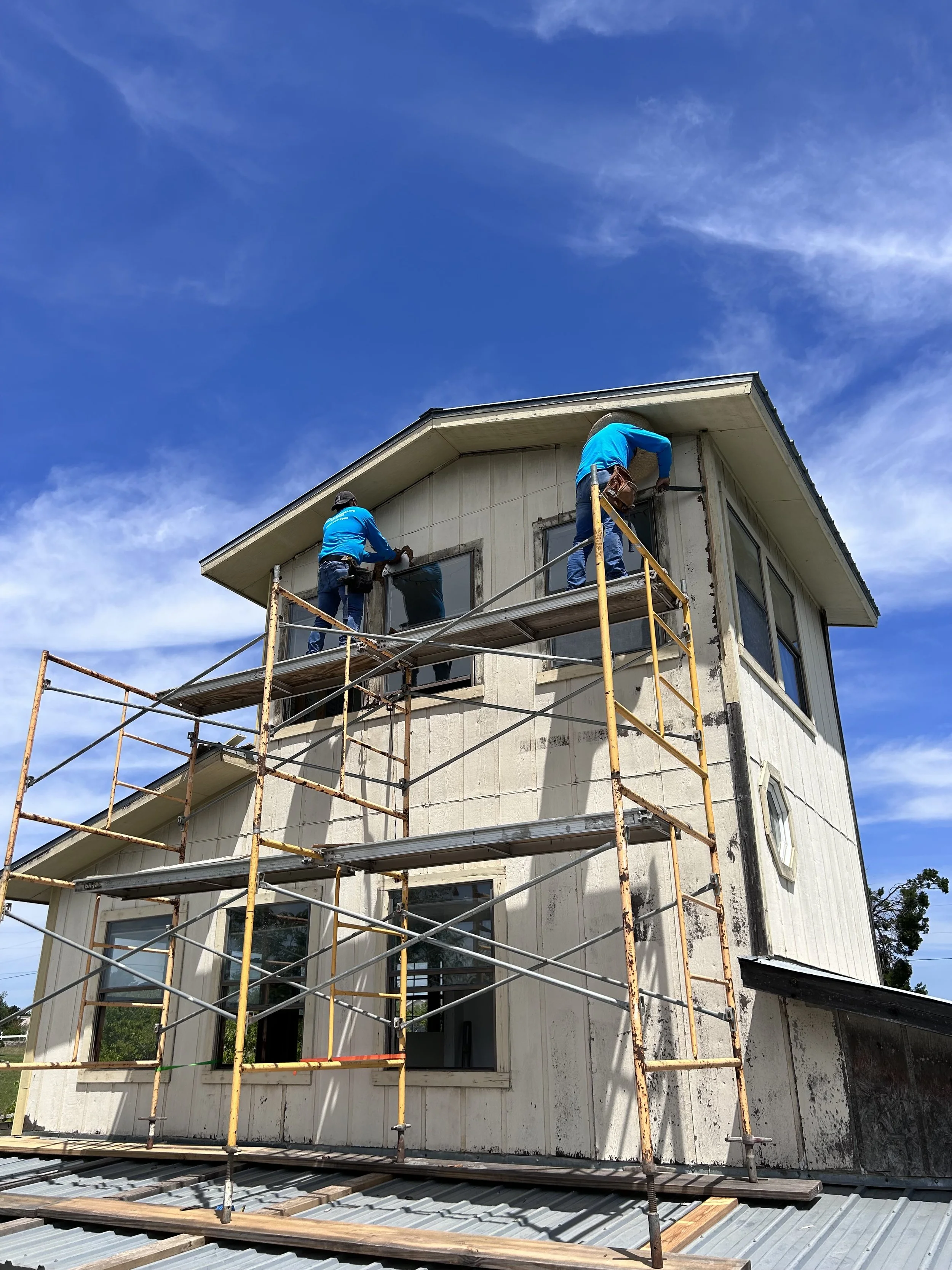 Two construction workers on scaffolding working on the exterior of a two-story house with peeling white siding under a blue sky.