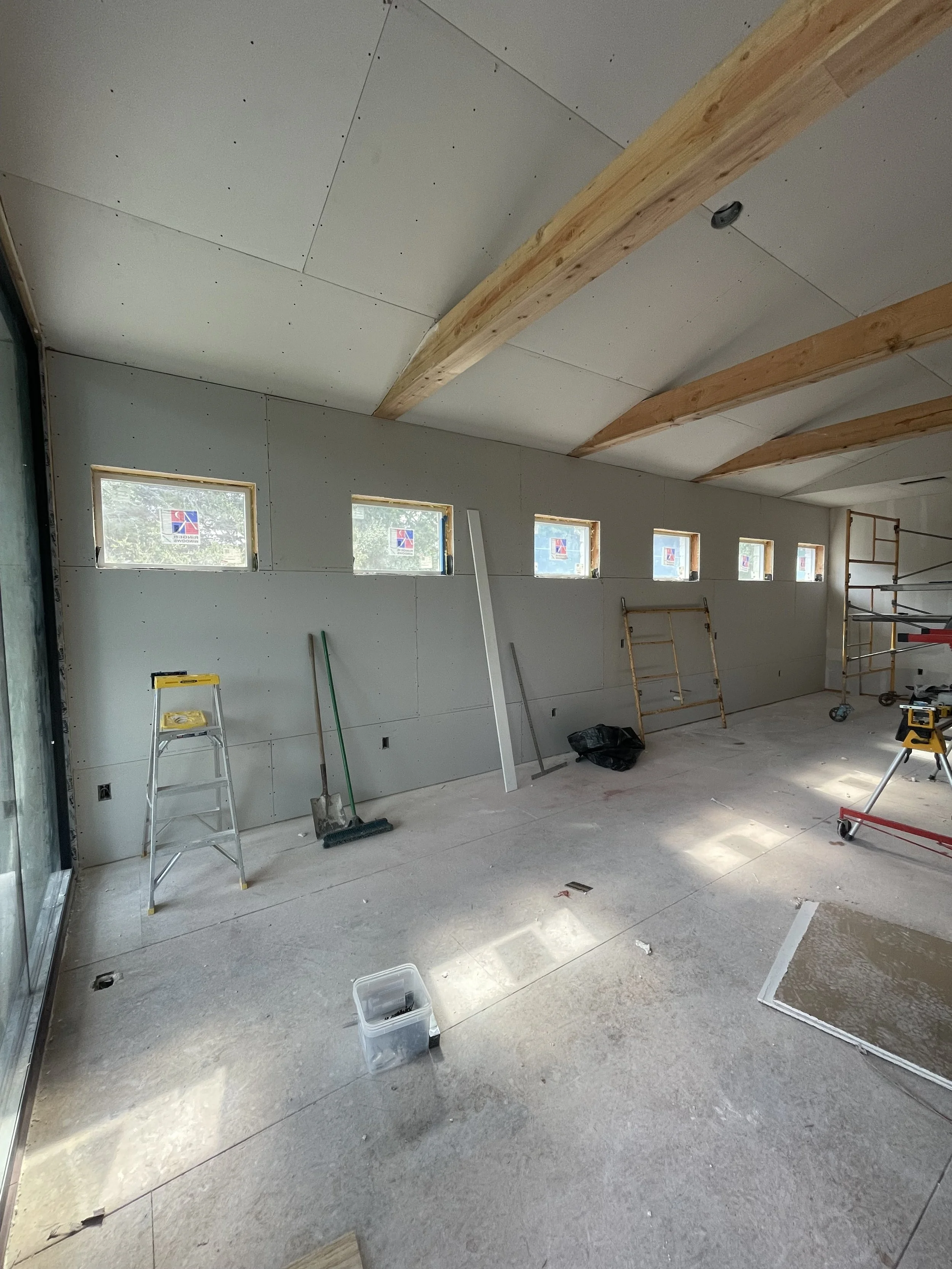 Interior of a room under construction with drywall installed on walls and ceiling, wooden beams on the ceiling, windows along the wall, and construction tools and materials scattered on the floor.
