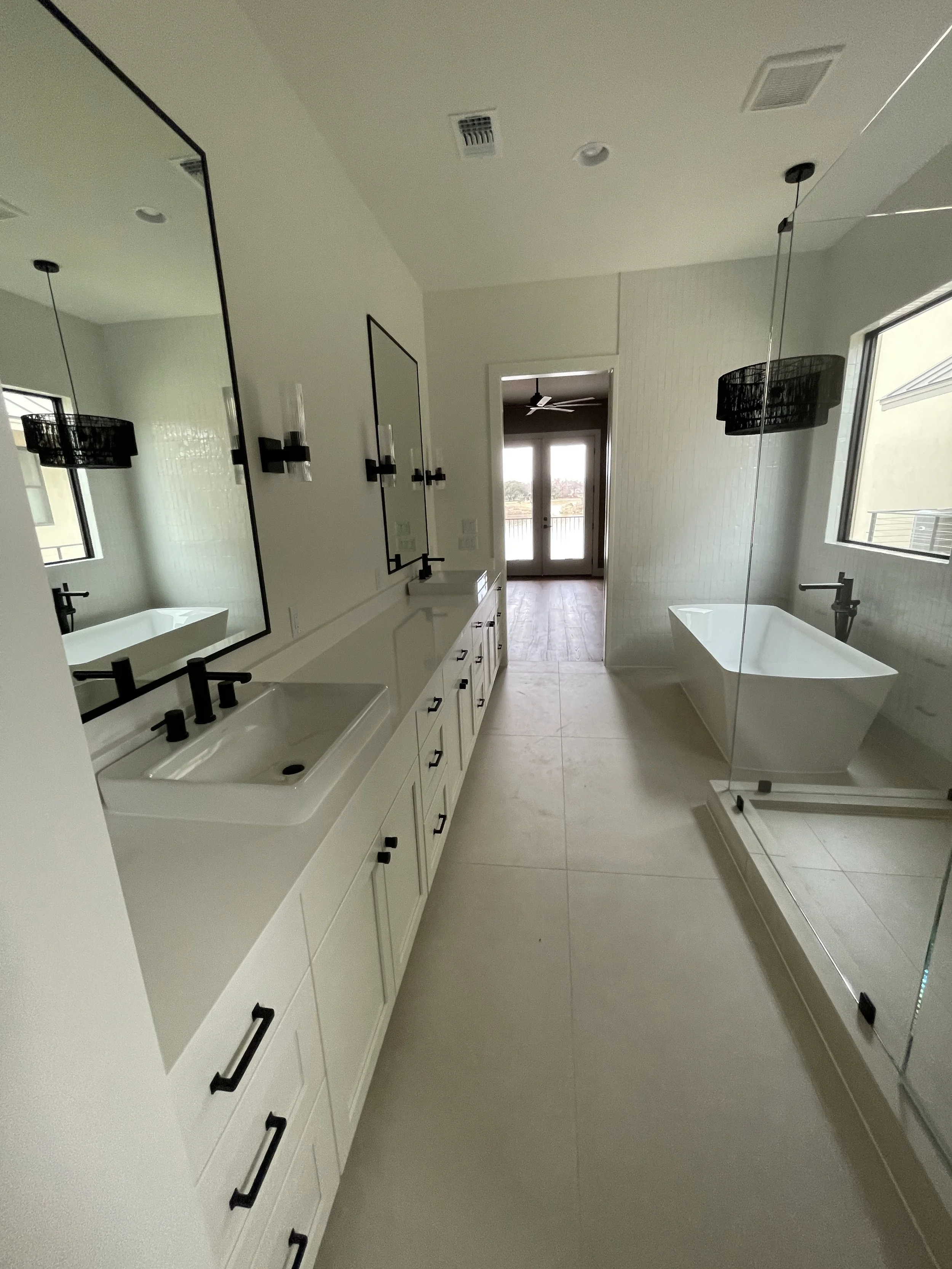 Modern white bathroom with double vanity, freestanding bathtub, black fixtures, pendant lights, and glass-enclosed shower.