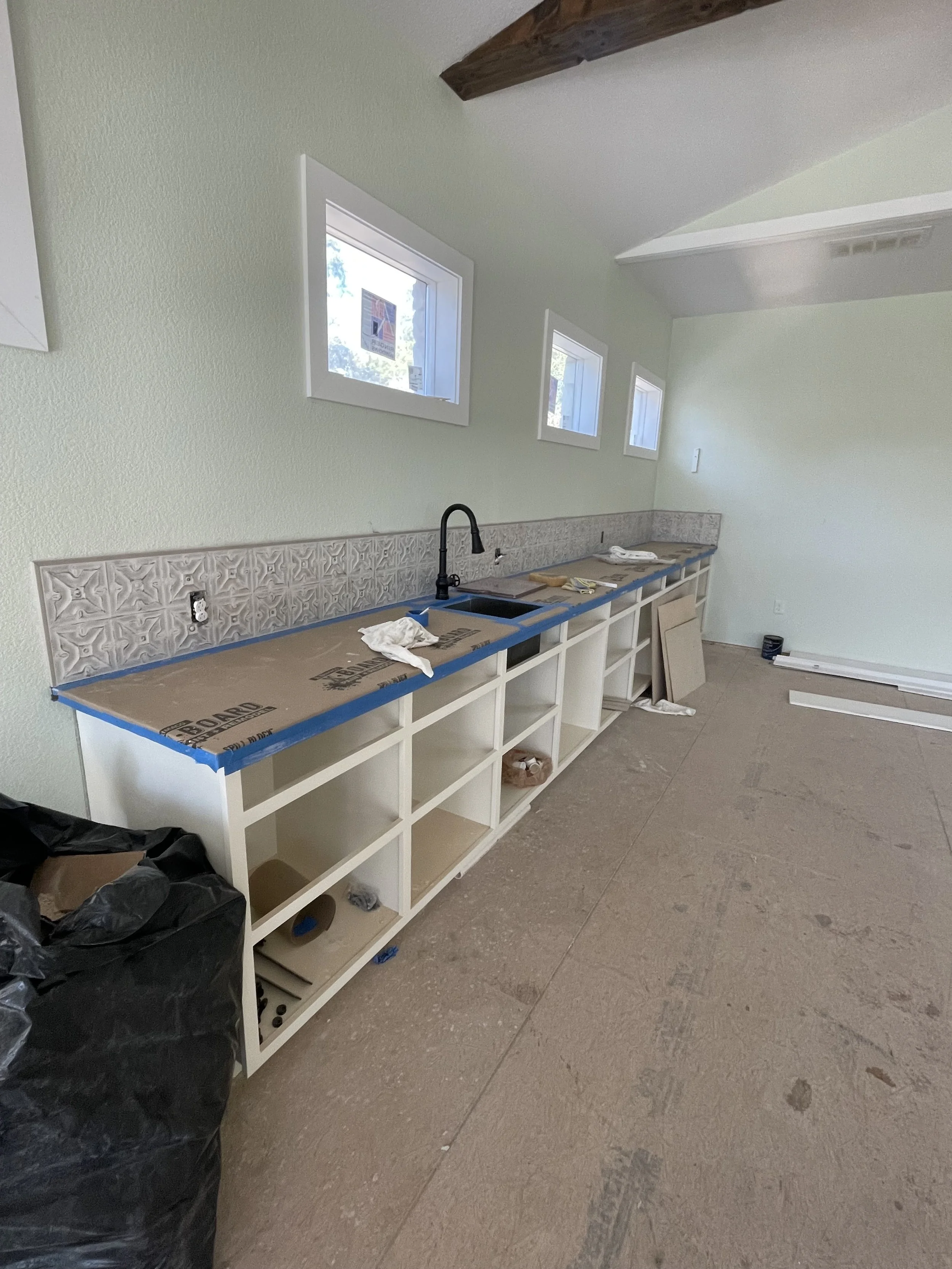 Under construction kitchen with light green walls, small windows, and an unfinished white cabinet with open shelves. A black faucet is installed above the sink area, and construction materials and tools are scattered on the counter and floor.
