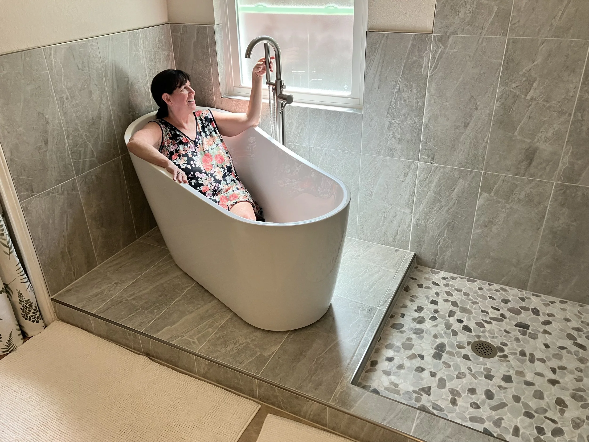 A woman with dark hair wearing a floral dress, sitting in a white freestanding bathtub in a tiled bathroom, holding the faucet handle and smiling.