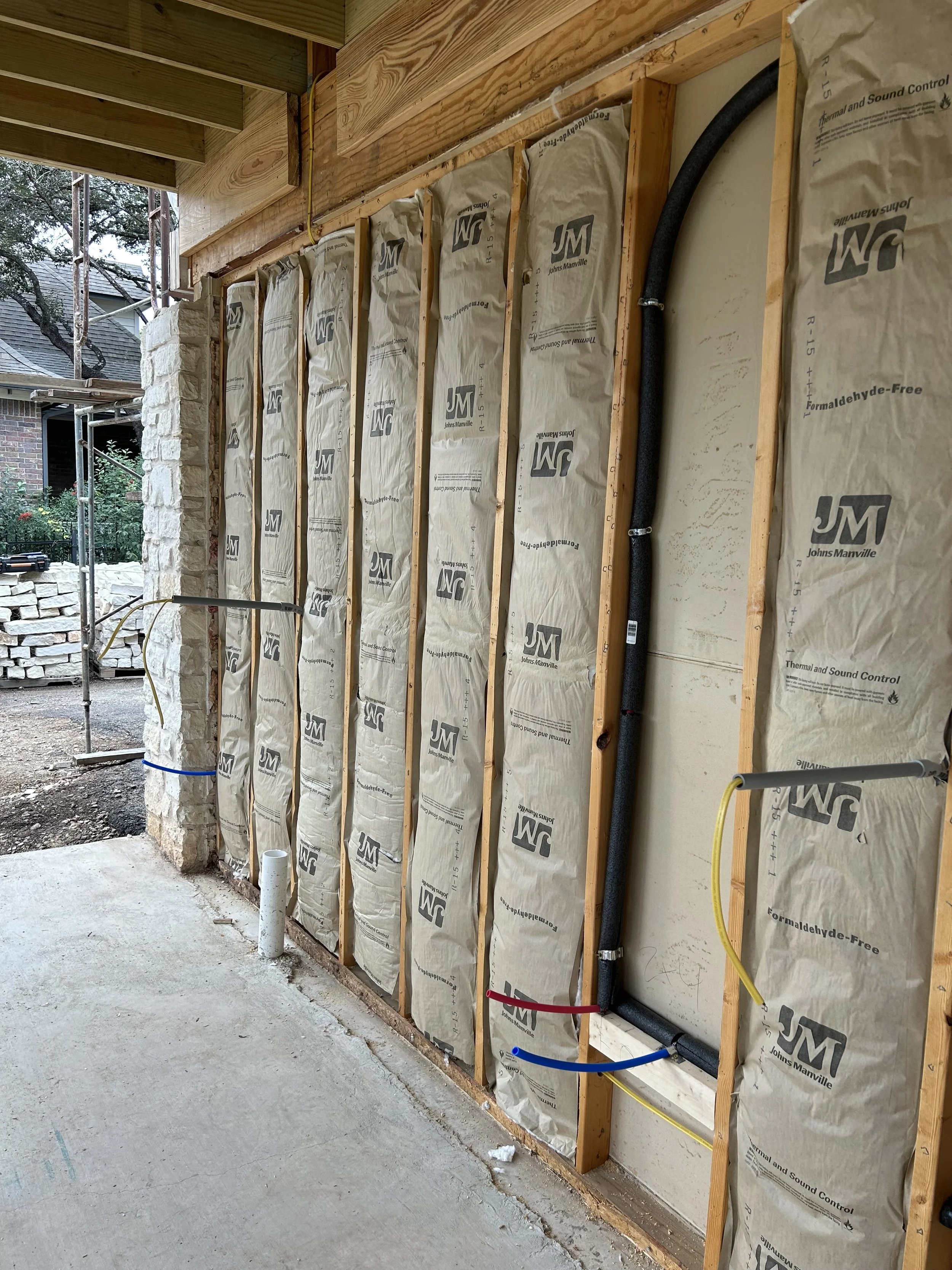 Partially constructed exterior wall with insulation, plumbing pipes, and electrical wiring visible. Wooden studs and framing are in place, with some papers and pipes installed.