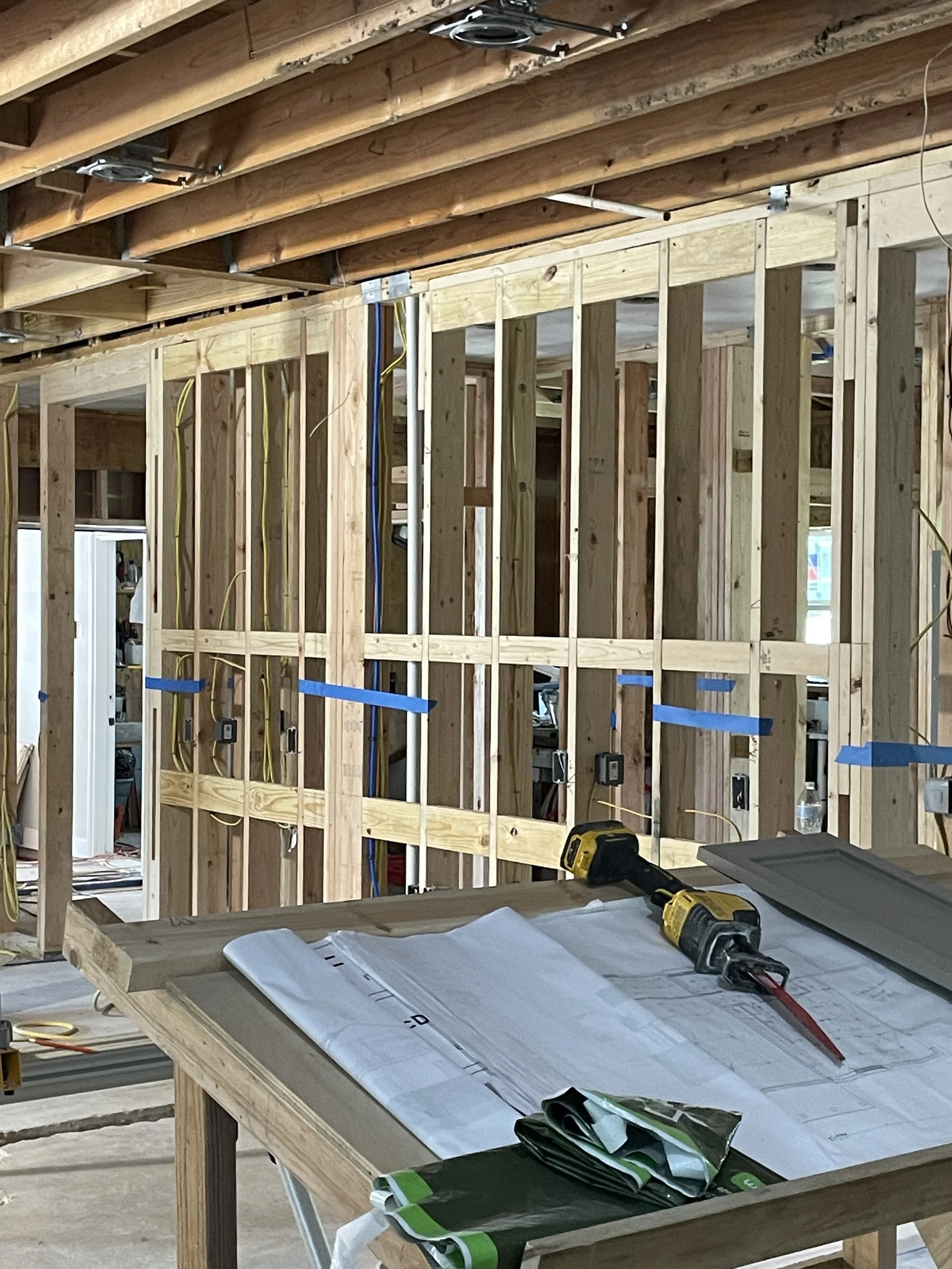 Interior of a construction site with exposed wooden framing, including studs and beams, with electrical wiring installed. A work table in the foreground holds construction plans, a cordless drill, and other tools and materials.