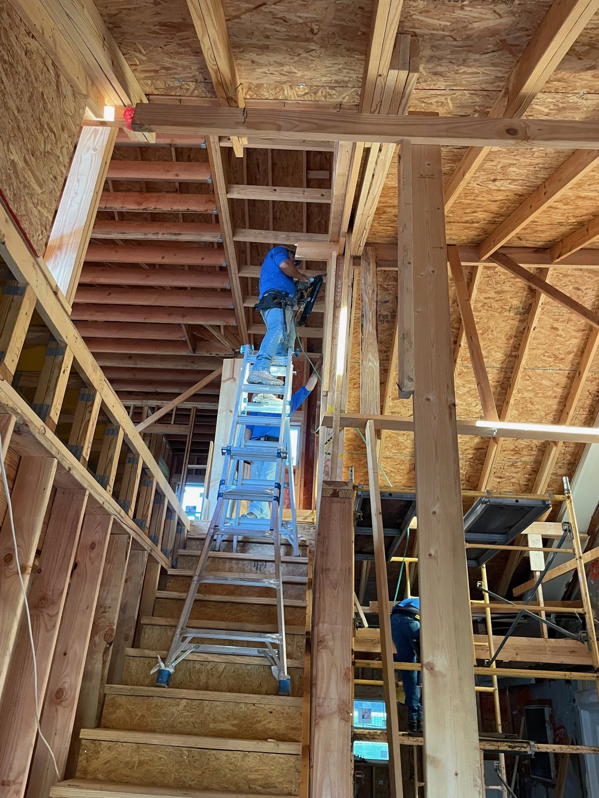 Construction workers on ladders inside a partially framed house with exposed wood studs, beams, and roof sheathing.