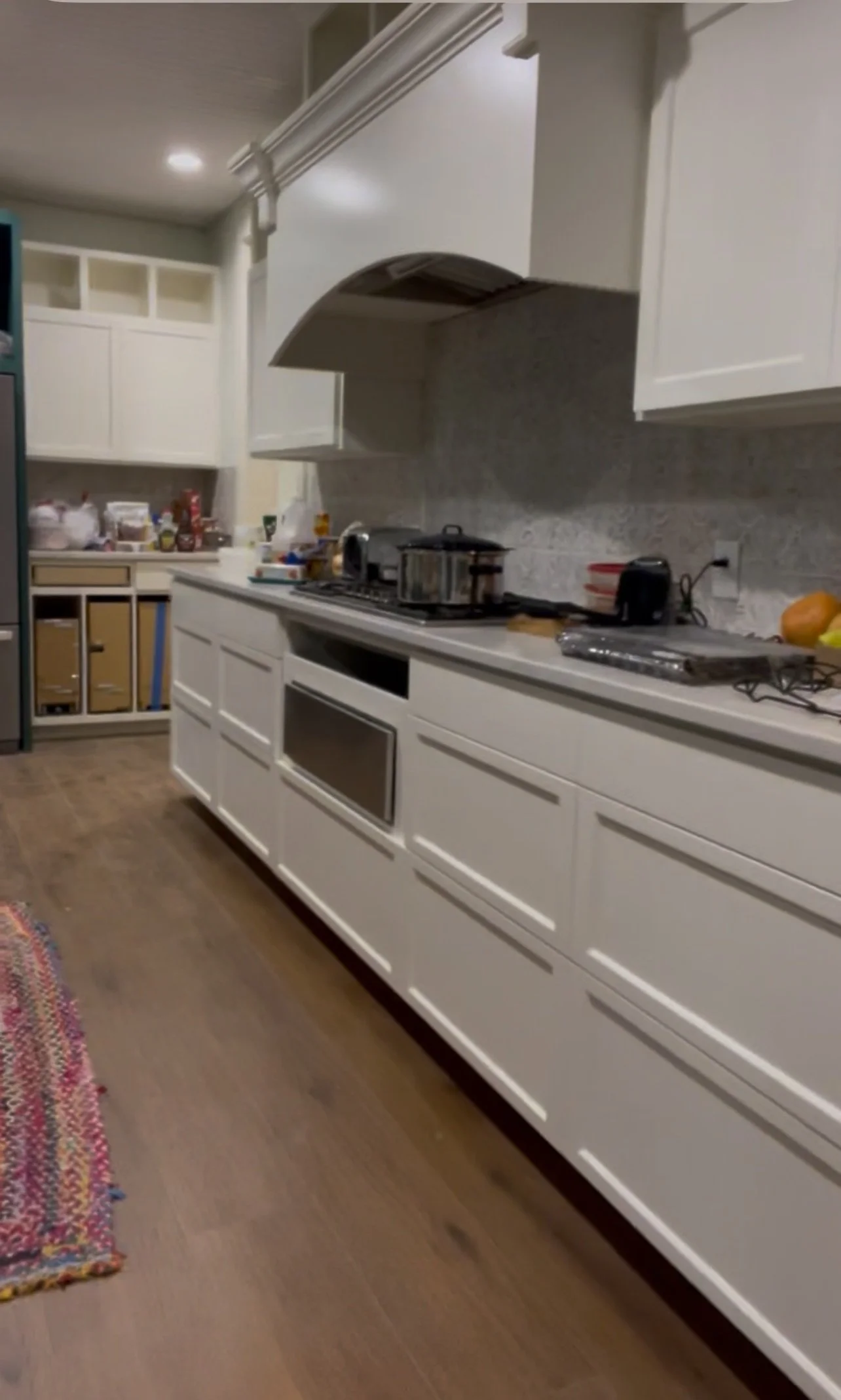 A kitchen with white cabinets, a marble countertop, and wooden flooring. Various appliances and items are on the counter, including a toaster, a microwave, and a pot on a stove.