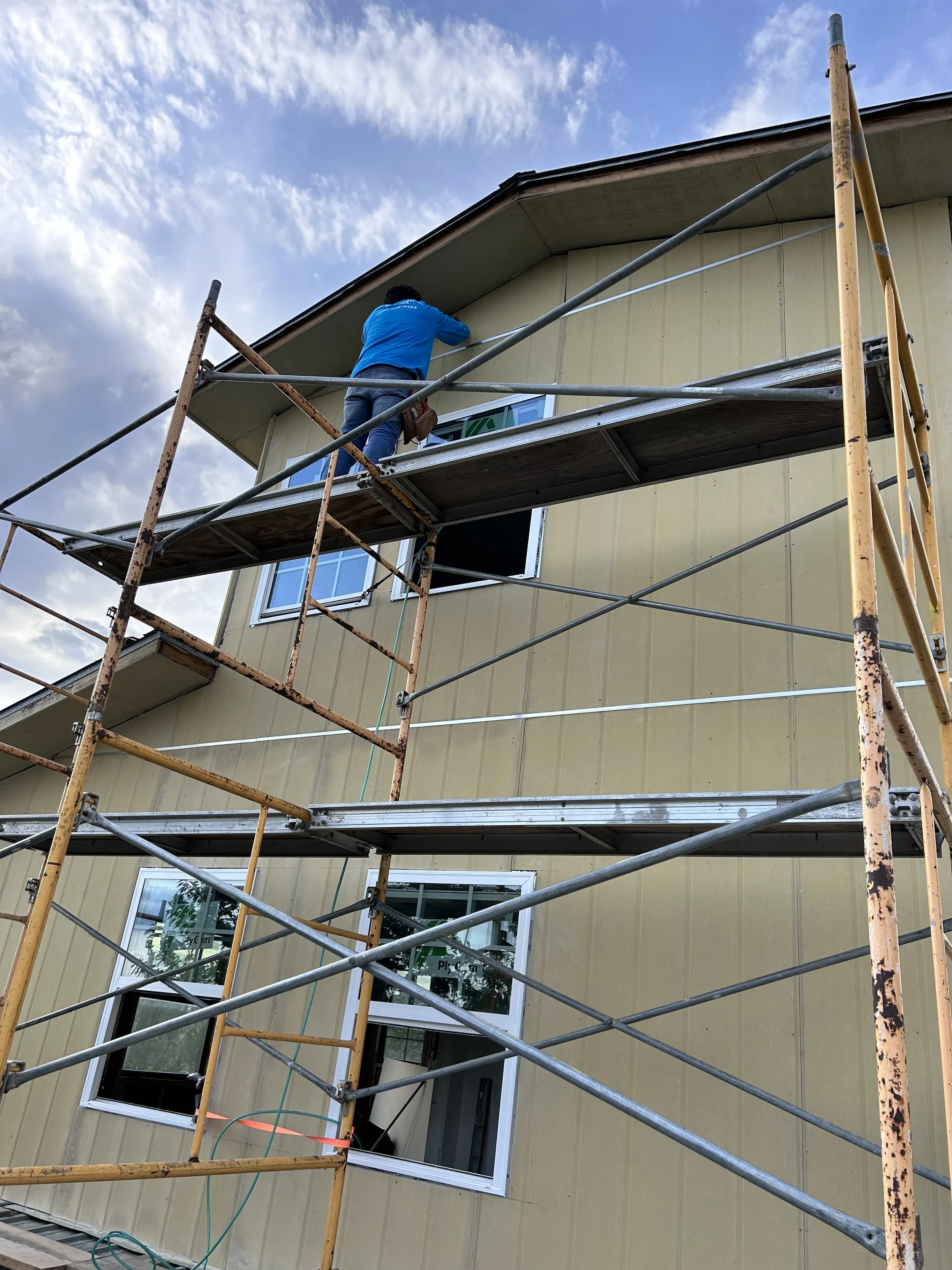 A person wearing a blue shirt and jeans working on a house roof from a scaffolding structure during construction or renovation.