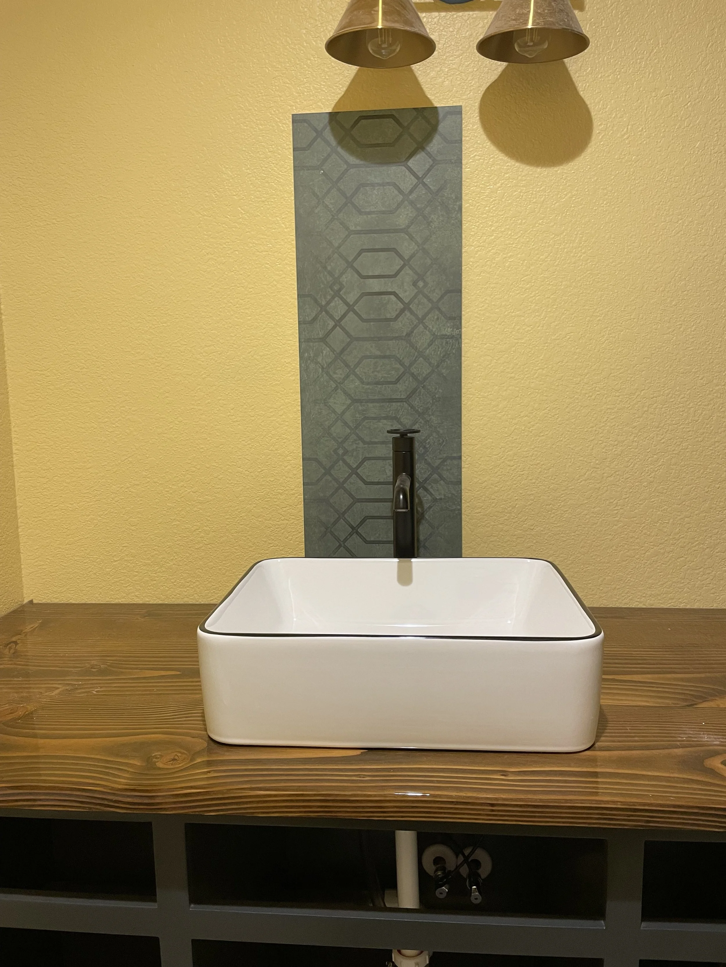 Bathroom sink with a black faucet on a wooden countertop, yellow wall background, two ceiling lights, and a patterned vertical tile behind the sink.