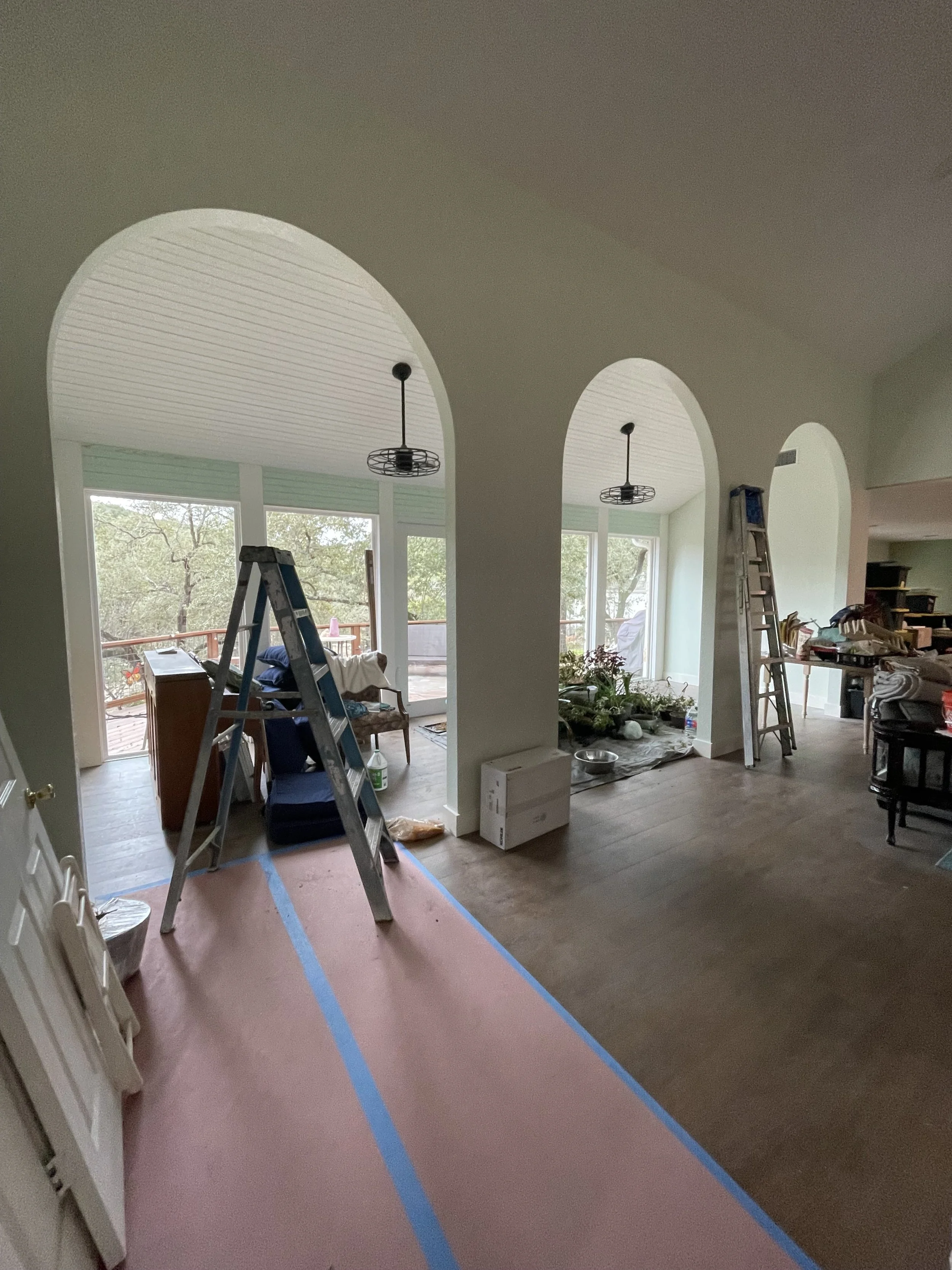 Living room and dining area under renovation, with ladders, construction materials, and blue painter's tape on the floor.