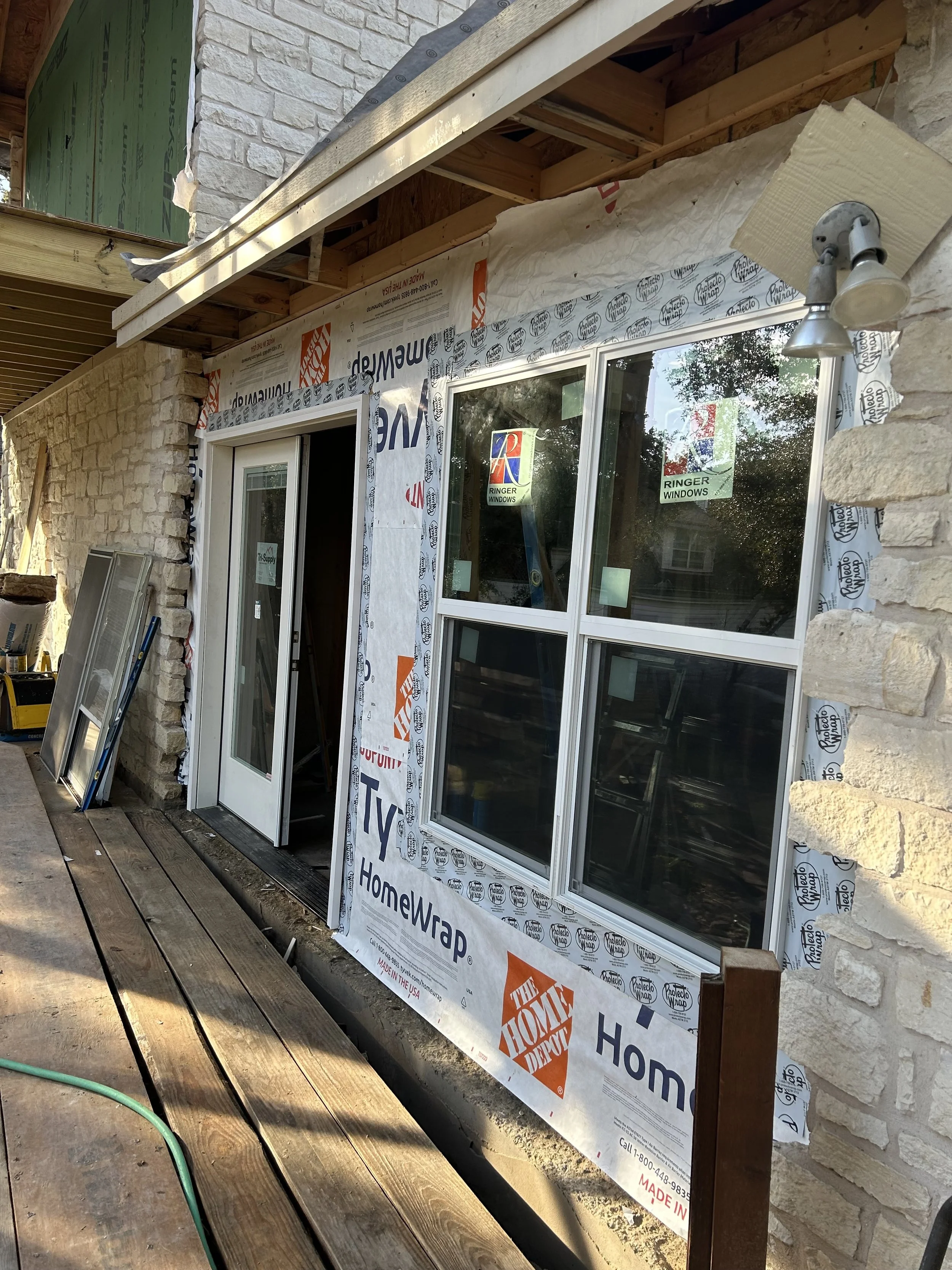 Construction site with a partially installed window and door, building materials, and a lamp fixture on a stone wall exterior.