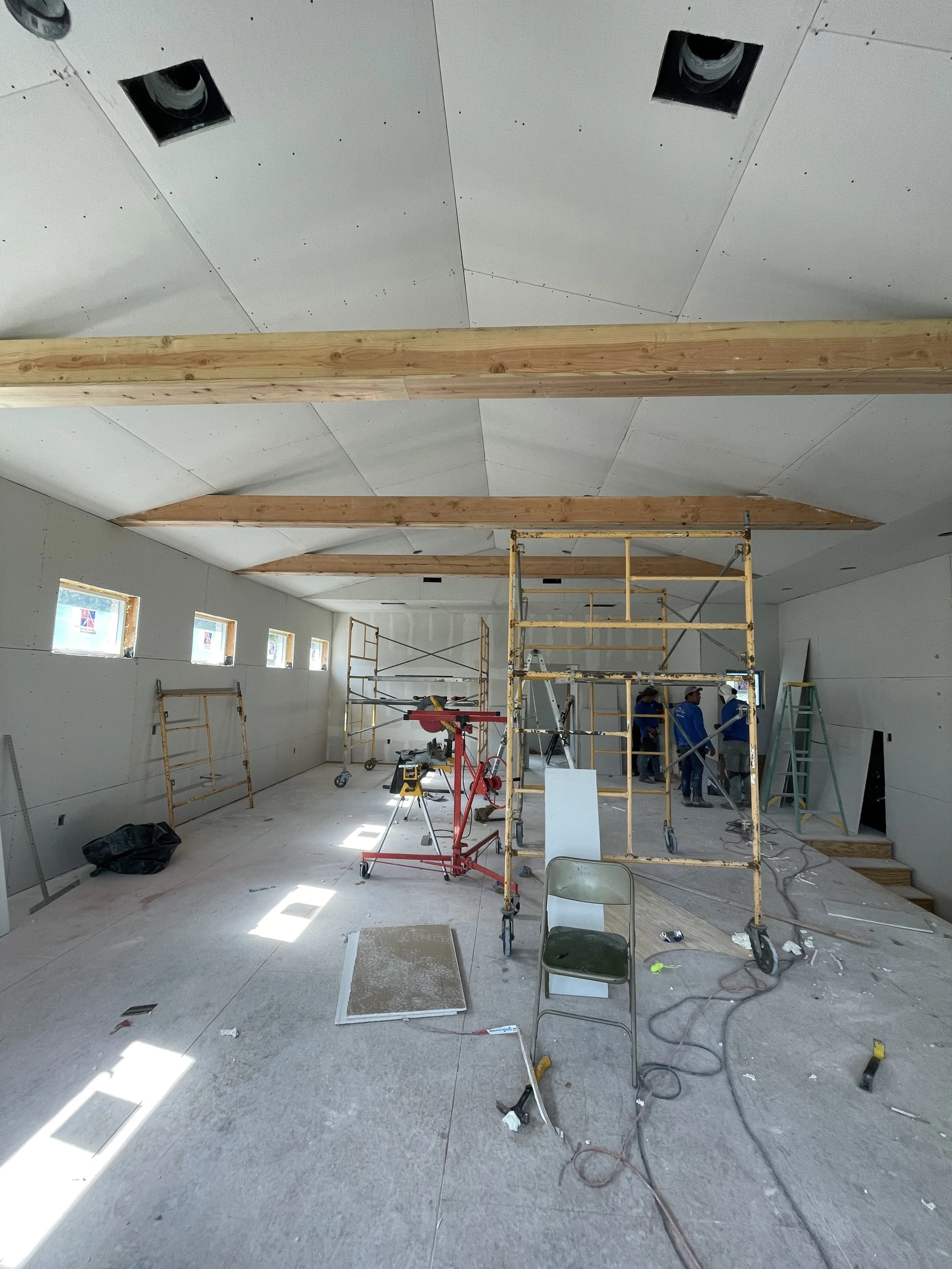 Interior of a room under construction with scaffolding, tools, and workers, ceiling with wooden beams, and small windows along one wall.